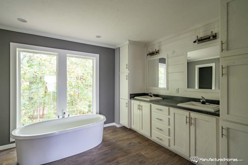 Modern bathroom with a large window, freestanding white tub, dual sinks with mirrors, and dark countertops. Features gray walls and wood flooring.