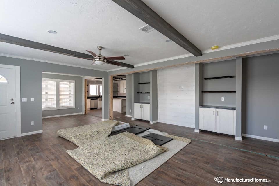 Spacious living room with gray walls, wooden floors, and exposed beams. Shelving units line one wall, with carpet padding in the center.