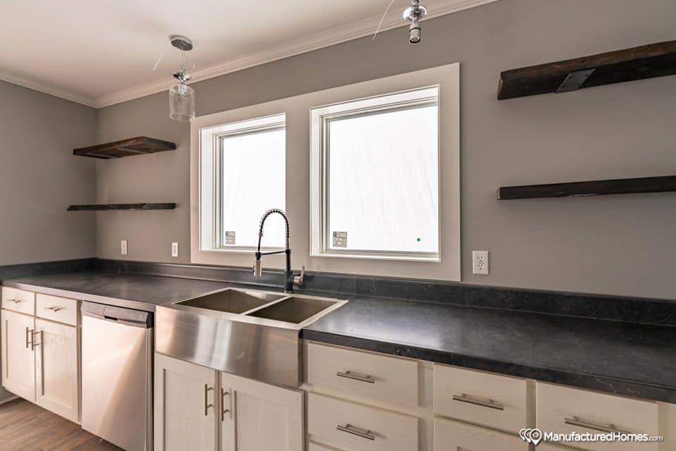 Modern kitchen with gray walls, featuring a large double sink and stainless steel faucet beneath two windows. Dark countertops contrast with white cabinets, and floating wooden shelves add minimalistic charm.