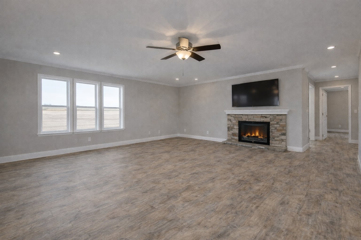 Spacious living room with wood floors, a ceiling fan, and a stone fireplace. Mounted TV above fireplace. Large windows let in natural light. Cozy and modern.