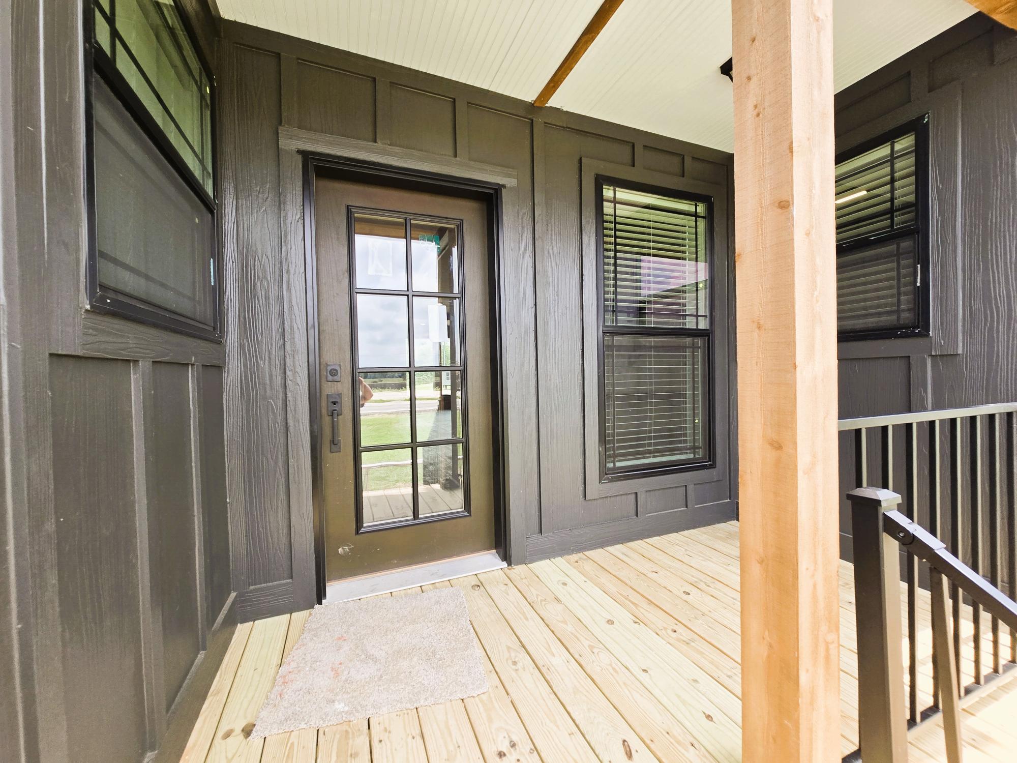 Wooden porch with a glass-panel door flanked by two large windows on a dark gray exterior wall. The scene conveys a welcoming and serene atmosphere.