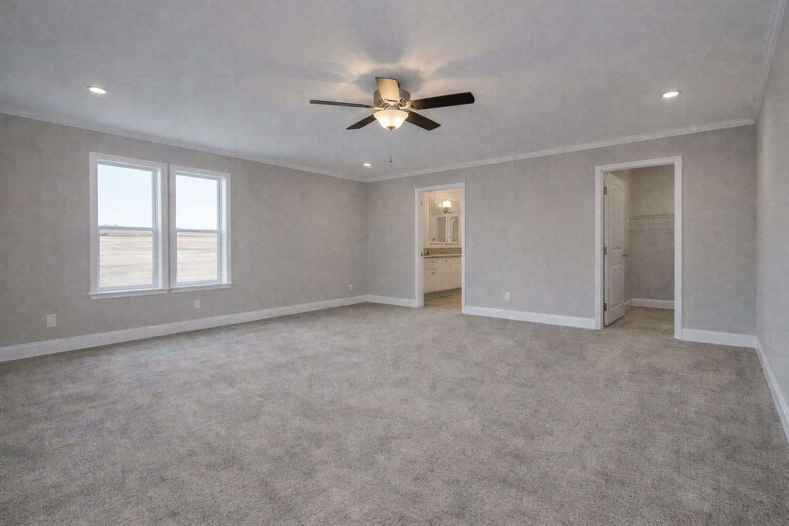 Spacious, empty bedroom with gray walls and carpet, a ceiling fan, and large windows. Open doors reveal a bathroom and closet, creating an inviting and serene atmosphere.