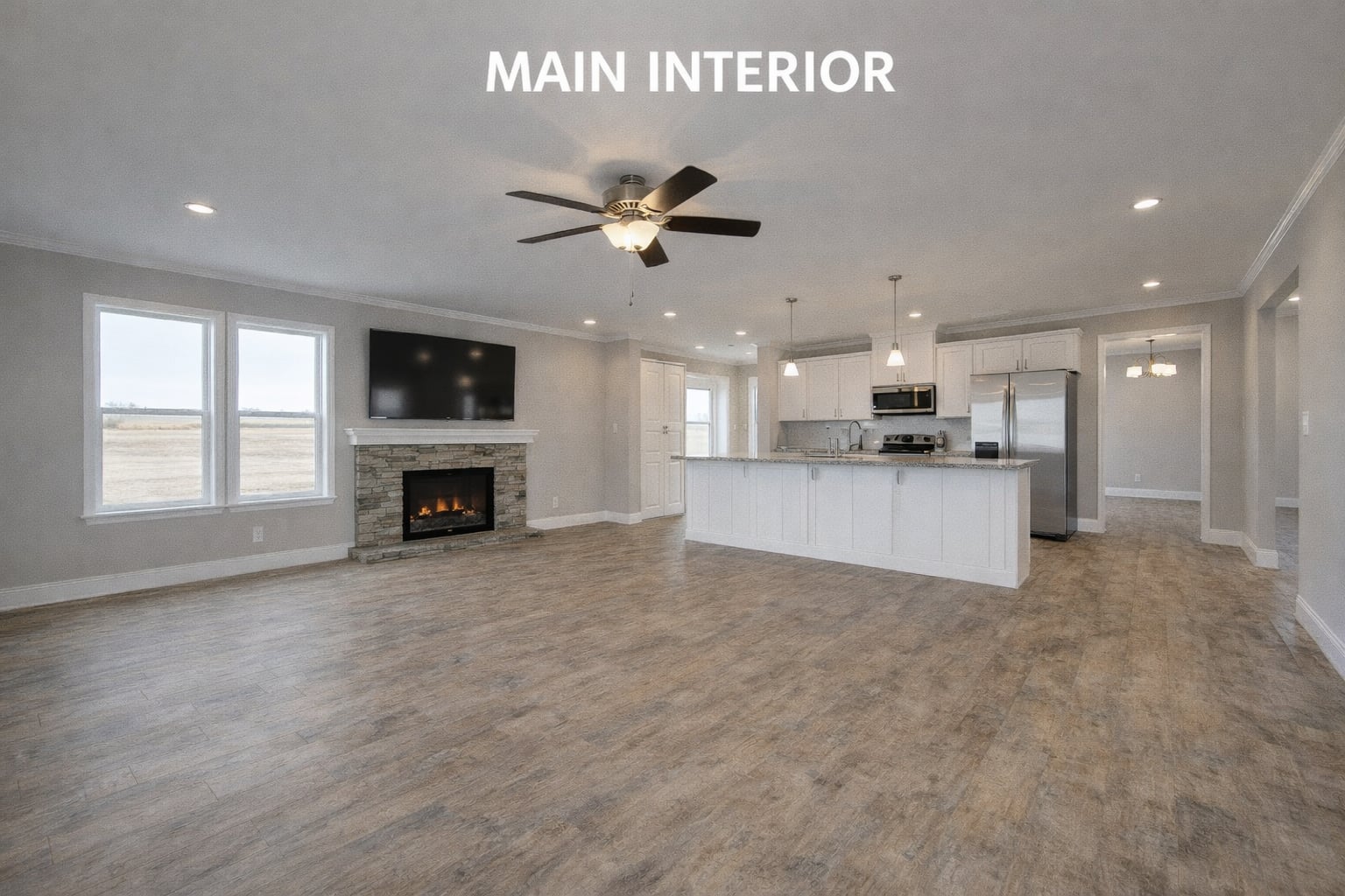 Spacious modern interior with wooden flooring, large kitchen island, stainless steel appliances, and a stone fireplace under a mounted TV. Ceiling fan and recessed lighting create a cozy ambiance.