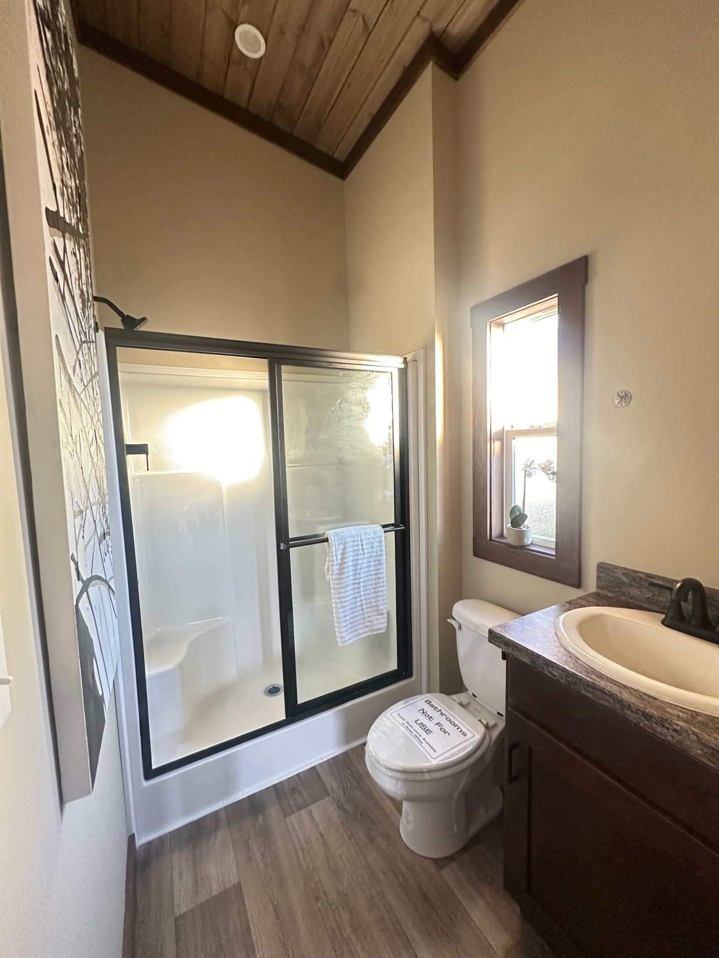 A bright bathroom with a wooden ceiling features a glass-enclosed shower and a toilet with a sign. Sunlight streams through a window above a dark wood vanity.