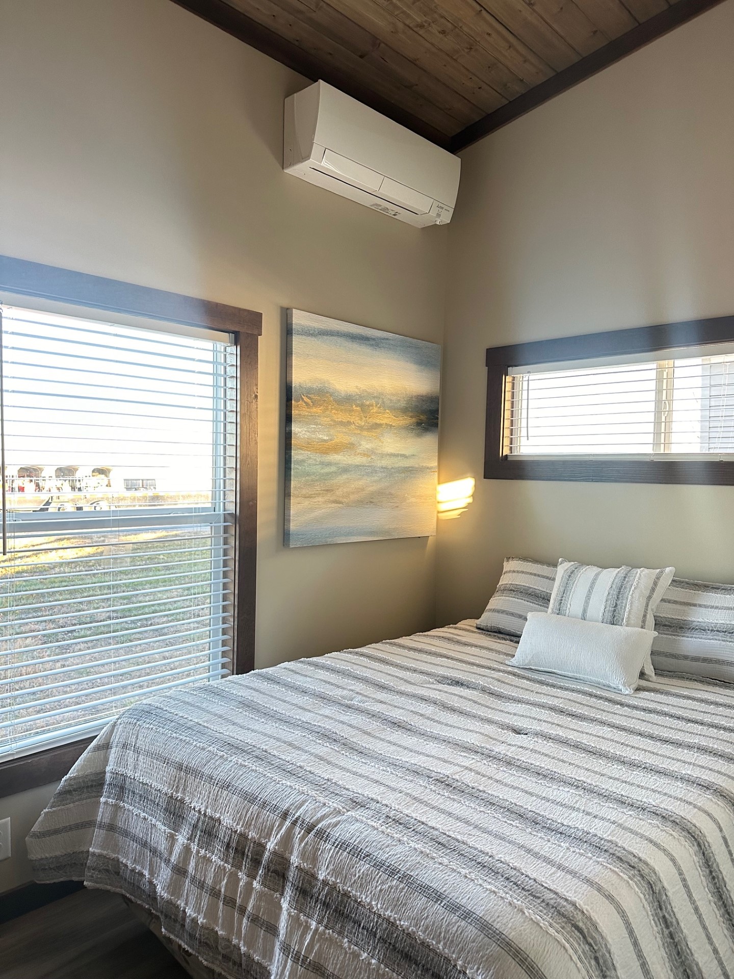 Cozy bedroom with a striped bedspread, abstract ocean painting, and soft natural light through two windows. Air conditioner above bed adds modern touch.