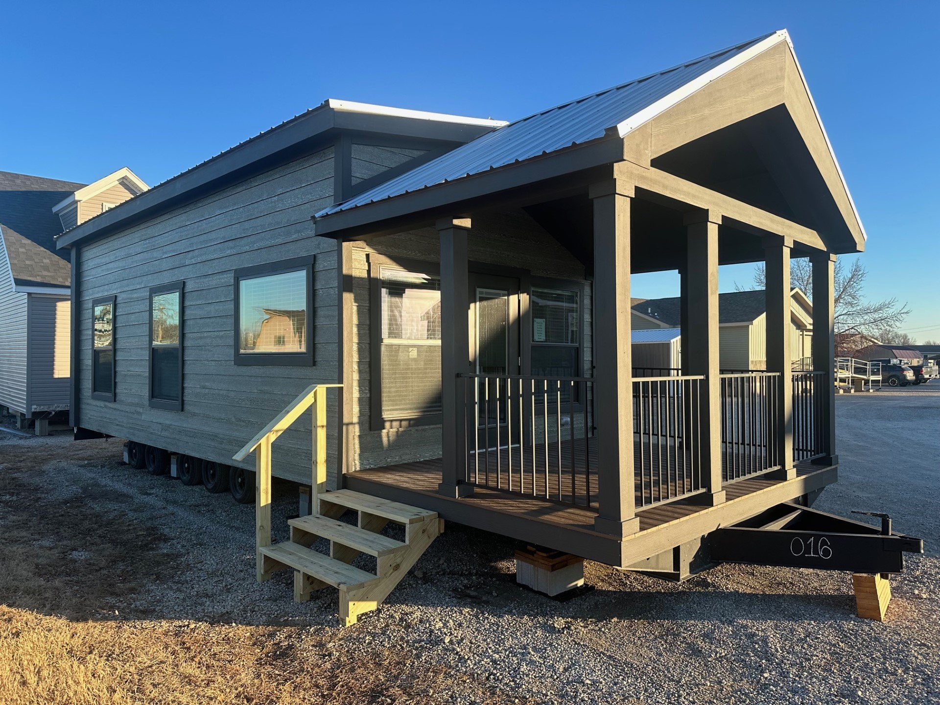 A modern tiny home with dark wood siding and a metal roof sits on gravel. It features a covered porch with railing and wooden steps, under a clear sky.
