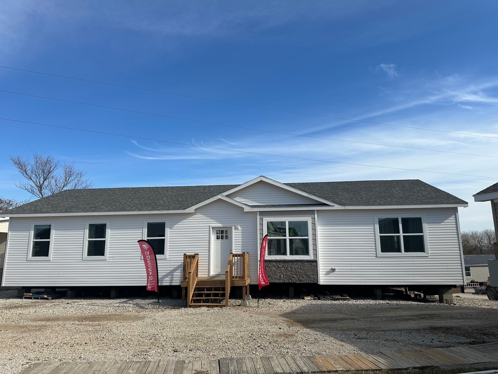 Single-story modular home with white siding, gray roof, and two red flags near wooden steps in a gravel area under a clear blue sky.