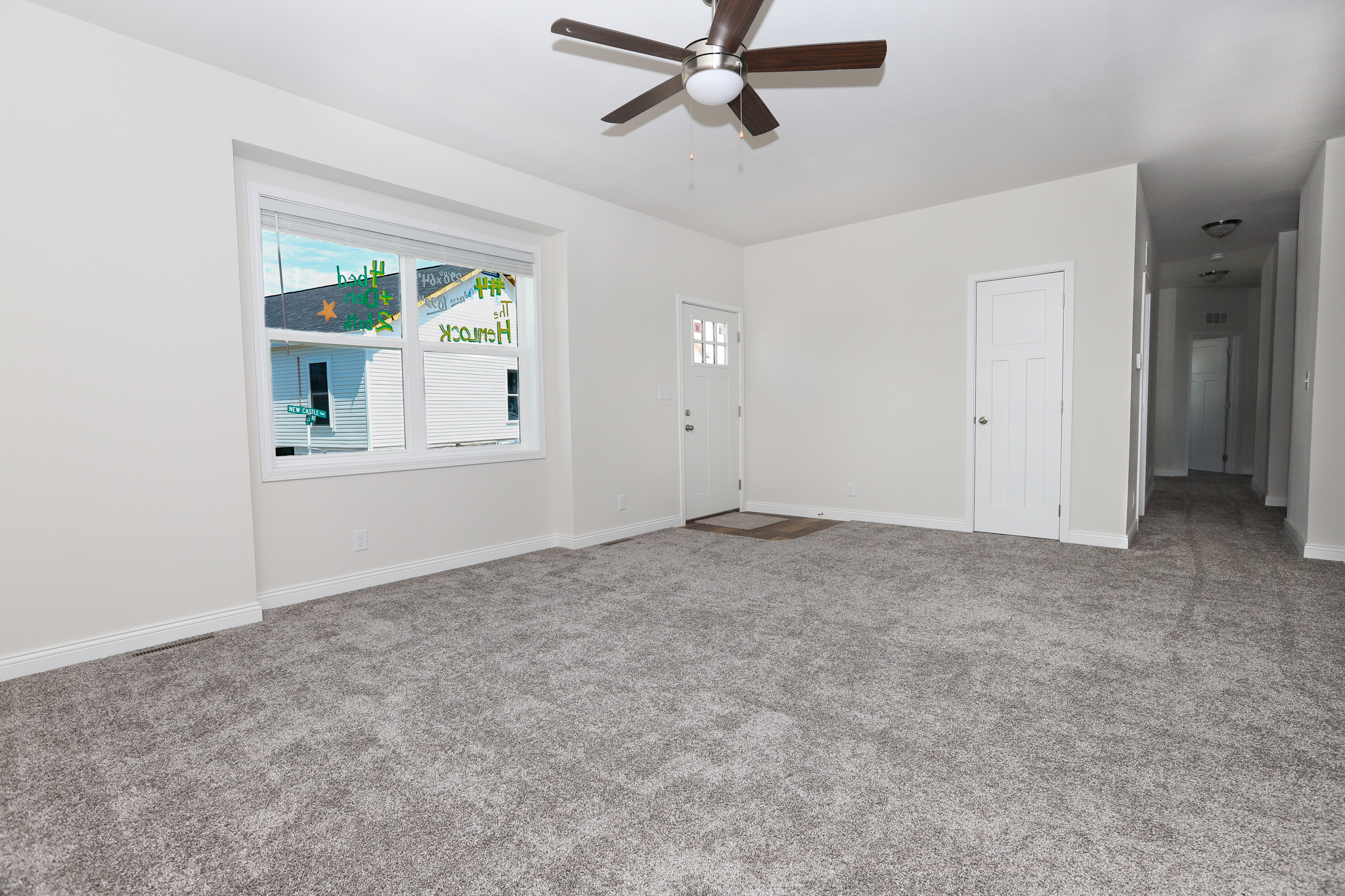 Empty living room with light gray carpet, white walls, and a ceiling fan. Large window and door on the left, a corridor leading to other rooms on the right.