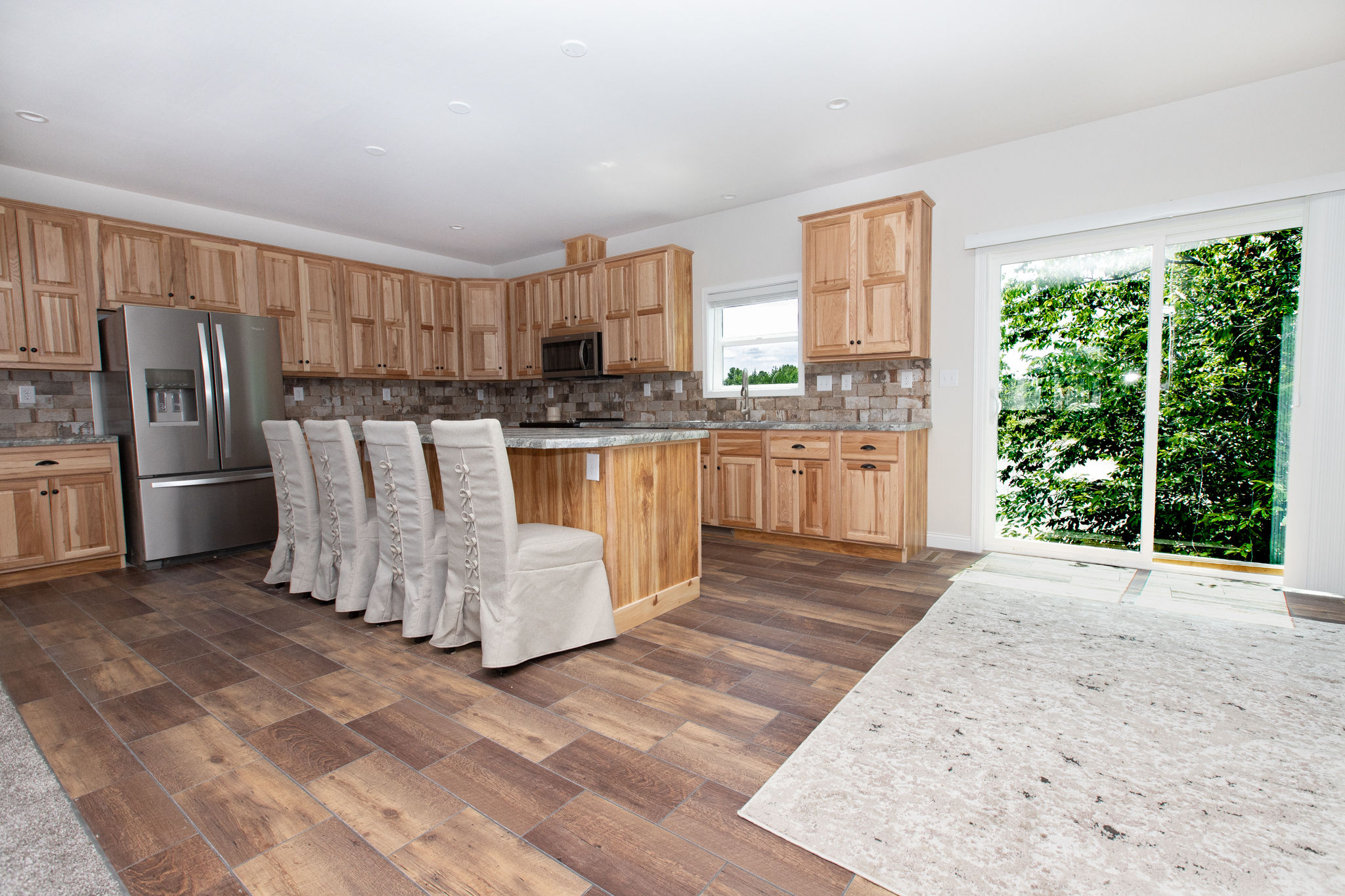 Spacious kitchen with light wood cabinets, island with beige slipcovered chairs, stainless steel appliances, and large windowed doors showing green foliage.