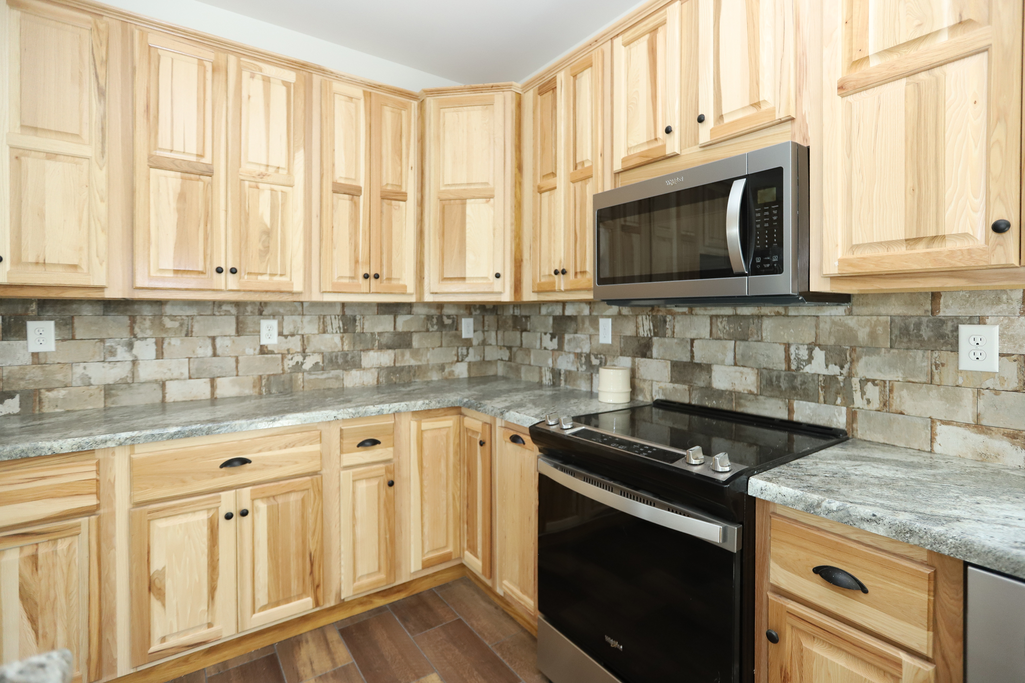 Modern kitchen with natural wood cabinets, granite countertops, and stone tile backsplash. Includes a stainless steel oven and microwave. Warm, rustic feel.