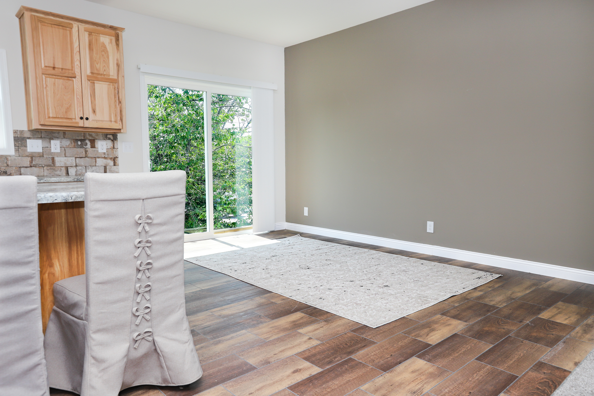 A bright, empty room with wooden floors, beige walls, and a large sliding glass door opening to greenery. Simple, modern, and inviting atmosphere.
