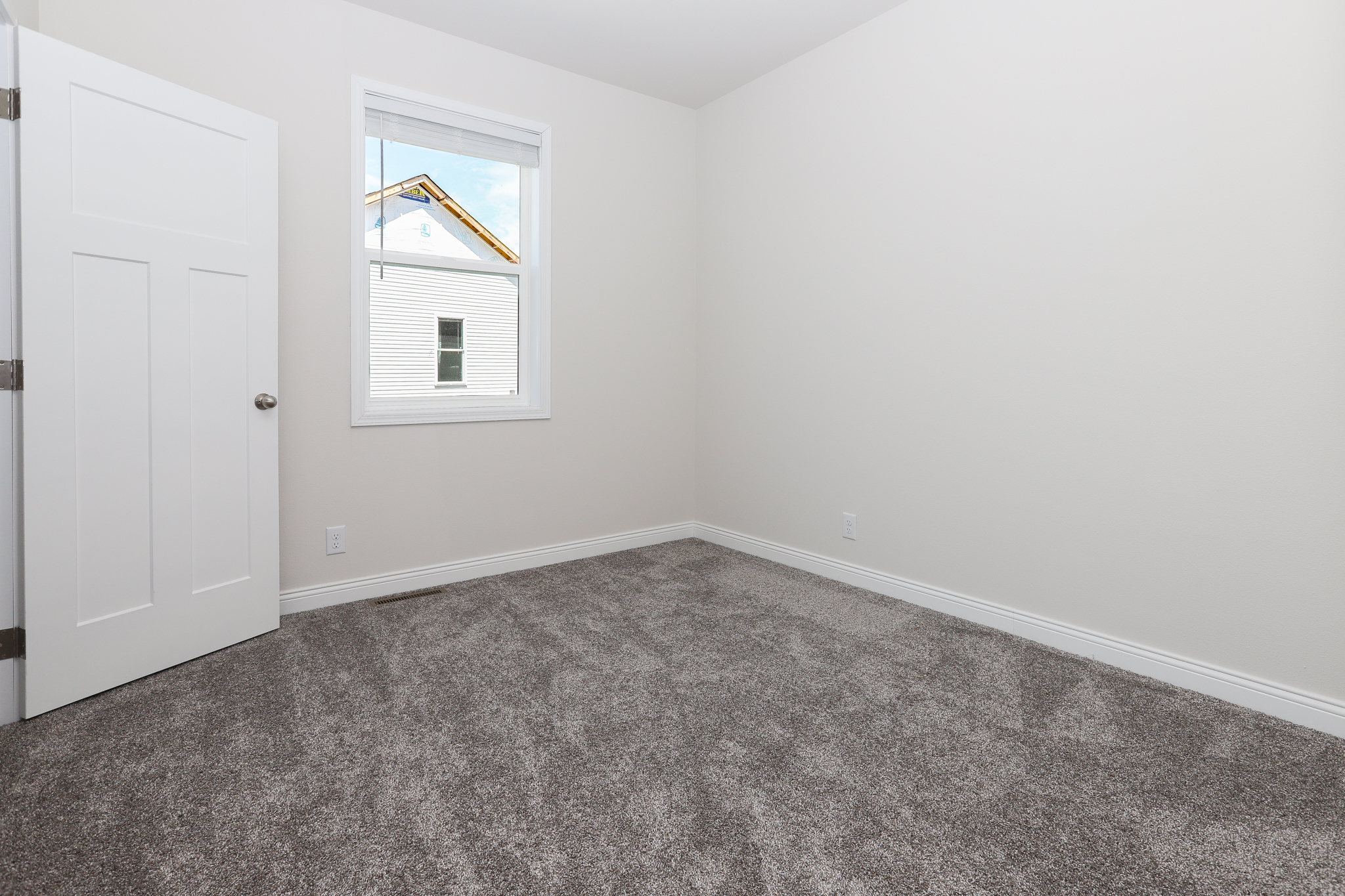 An empty, bright room with white walls and a gray carpeted floor. A single window shows a neighboring building. A closed white door is on the left.