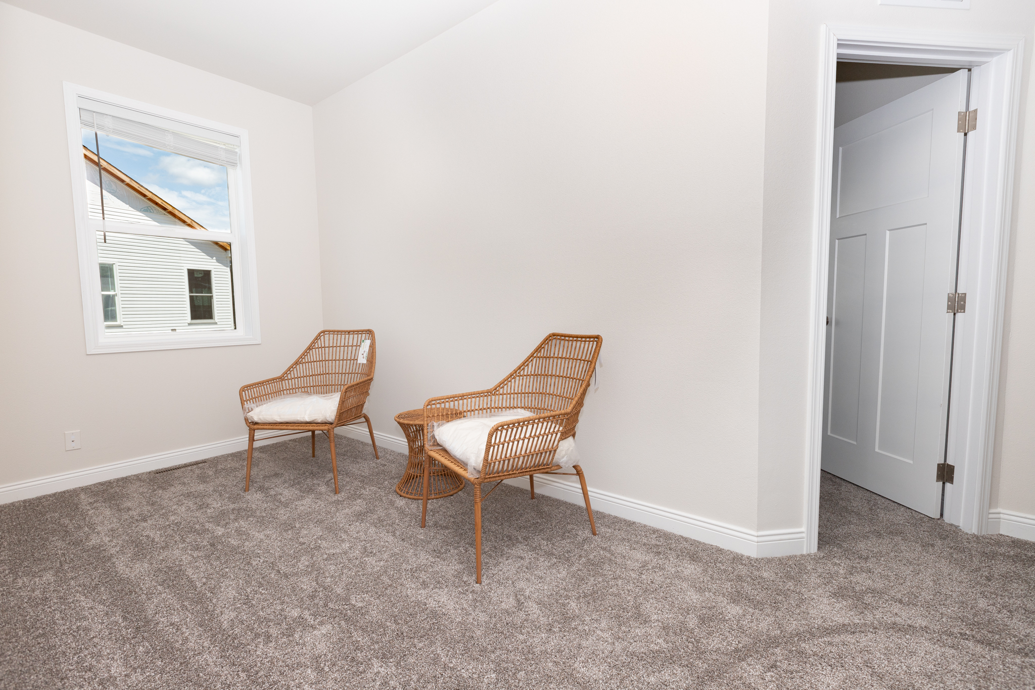 Two wicker chairs with cushions face each other in a small, carpeted room with white walls. A window shows blue sky and a neighboring house's roof. Calm and minimalist.