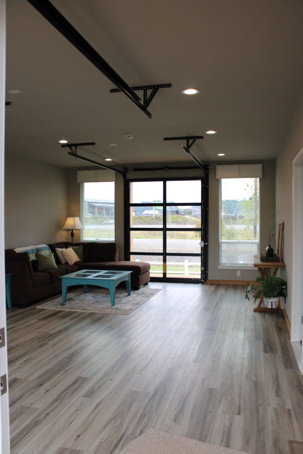 Modern living room with light wood flooring, brown sectional sofa, and turquoise coffee table. Large windows with natural light and outside view. Cozy and spacious ambiance.