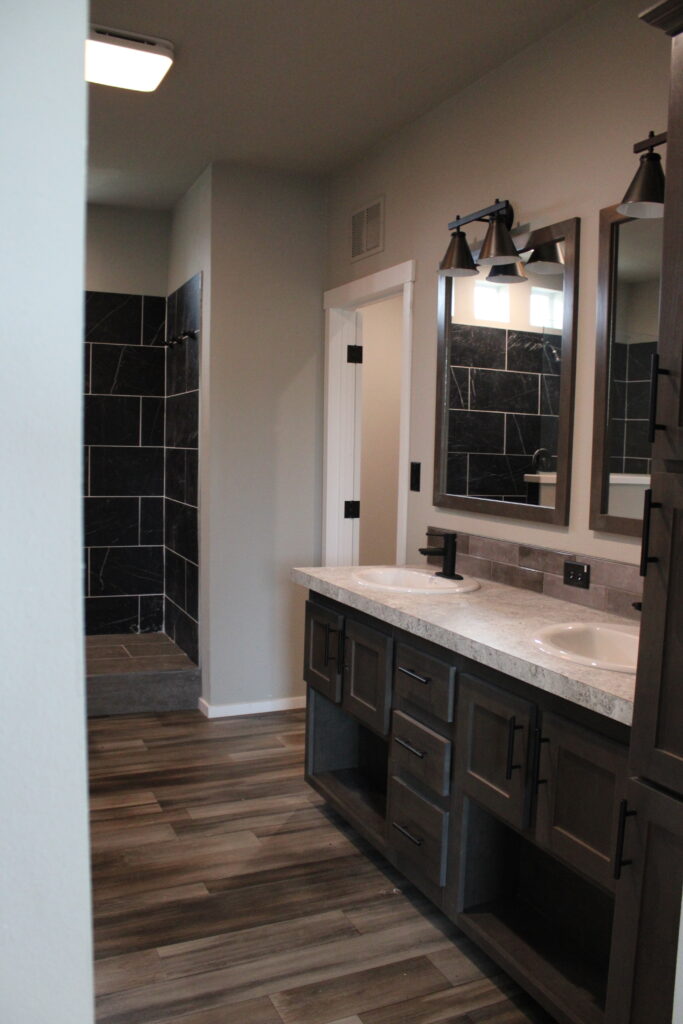 Modern bathroom with dark tile shower and dual-sink vanity. Wooden cabinets, sleek black fixtures, and warm lighting create a cozy and elegant atmosphere.