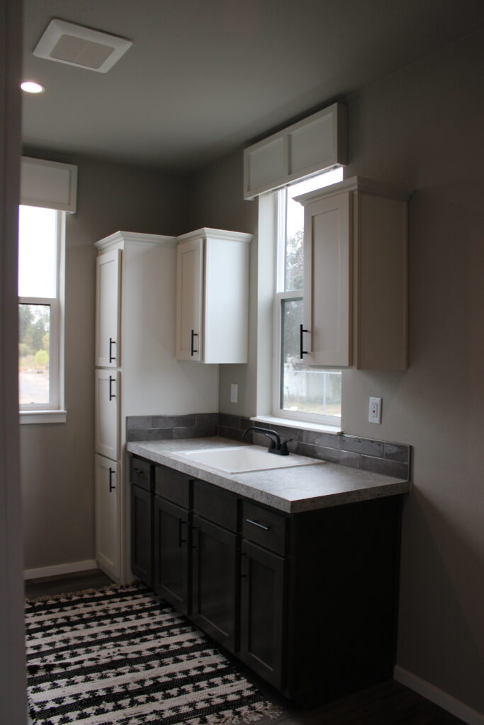 A modern kitchen corner with gray walls, dark wood lower cabinets, and white upper cabinets. Sunlight streams through the windows, highlighting a black and white patterned rug on the floor.