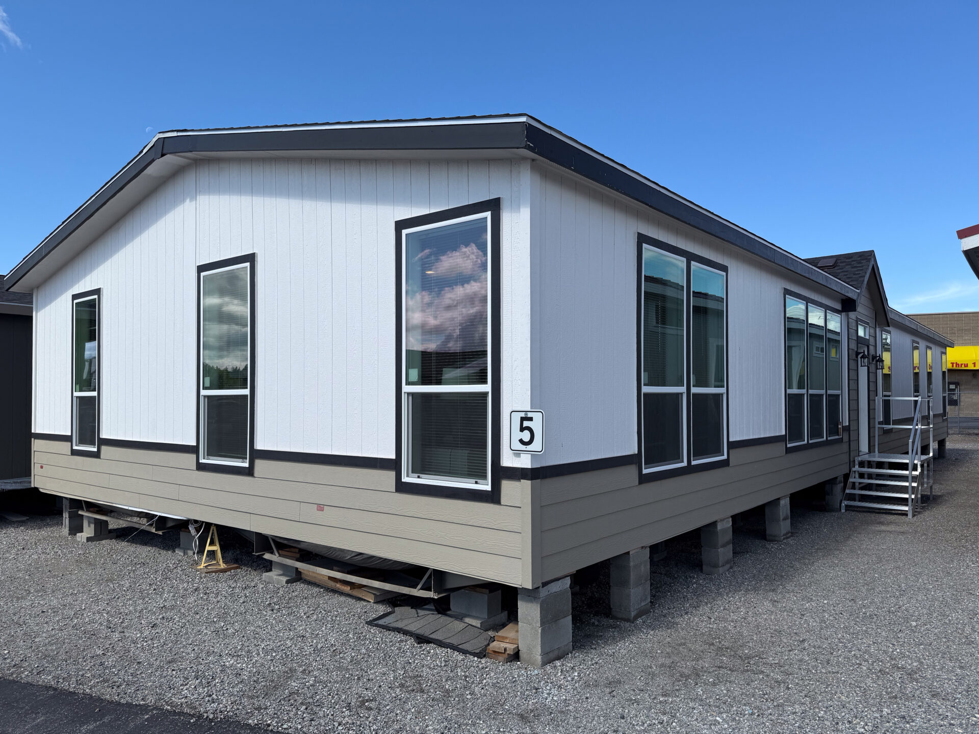 Single-story white and beige manufactured home elevated on concrete blocks. Large windows line the side, numbered "5", on a gravel lot under a clear blue sky.