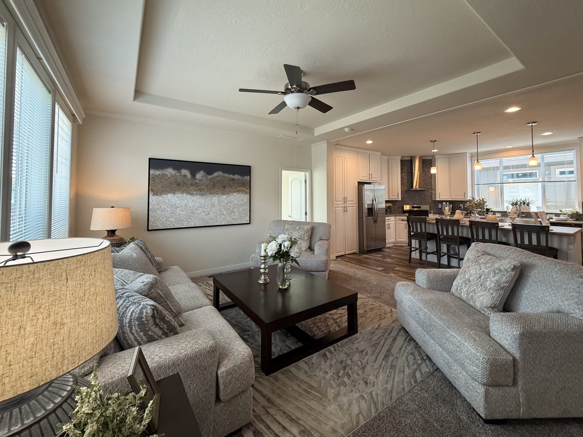 Modern living room with gray sofas, a dark wood coffee table, and a textured rug. Artwork adorns the wall. An open kitchen with a dining area, pendant lights, and bar stools connects to the space. A ceiling fan adds a touch of elegance.