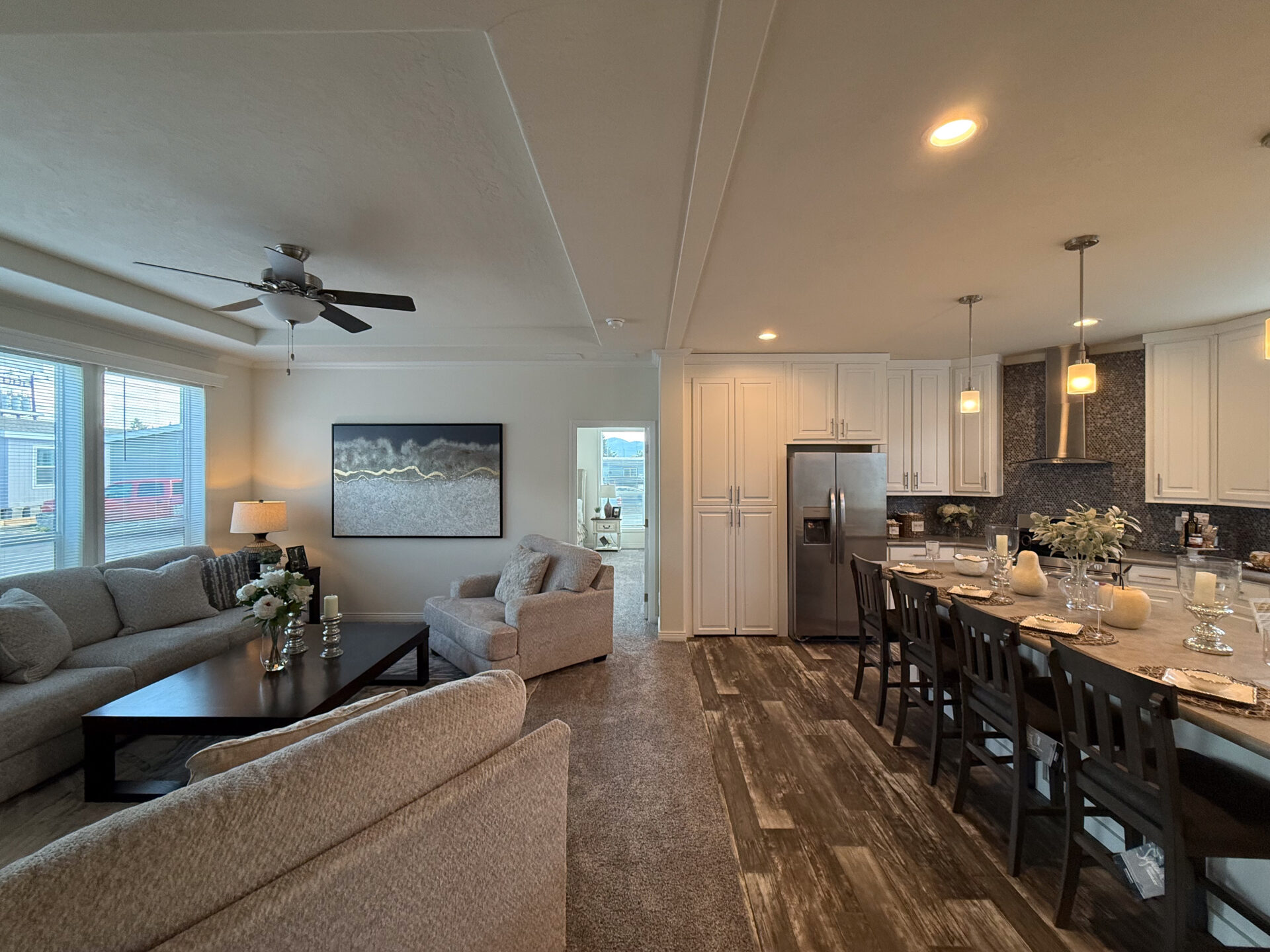 Modern living room and kitchen with gray sofas, a ceiling fan, and wall art. Kitchen features bar seating, pendant lights, and stainless steel appliances.
