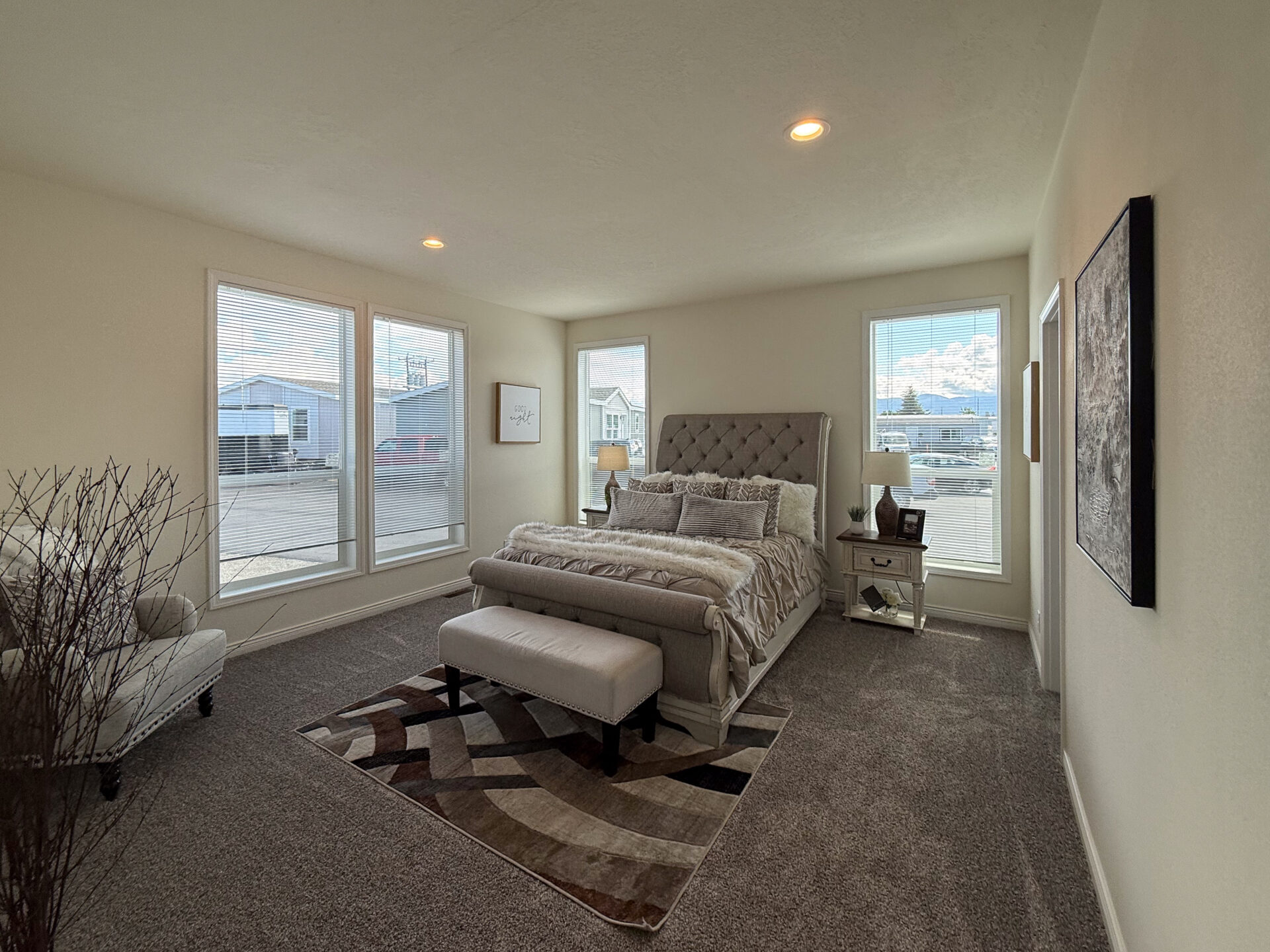 Cozy bedroom with a beige carpet and tufted bed. Large windows allow natural light, and decor includes a decorative rug, paintings, and a small bench.