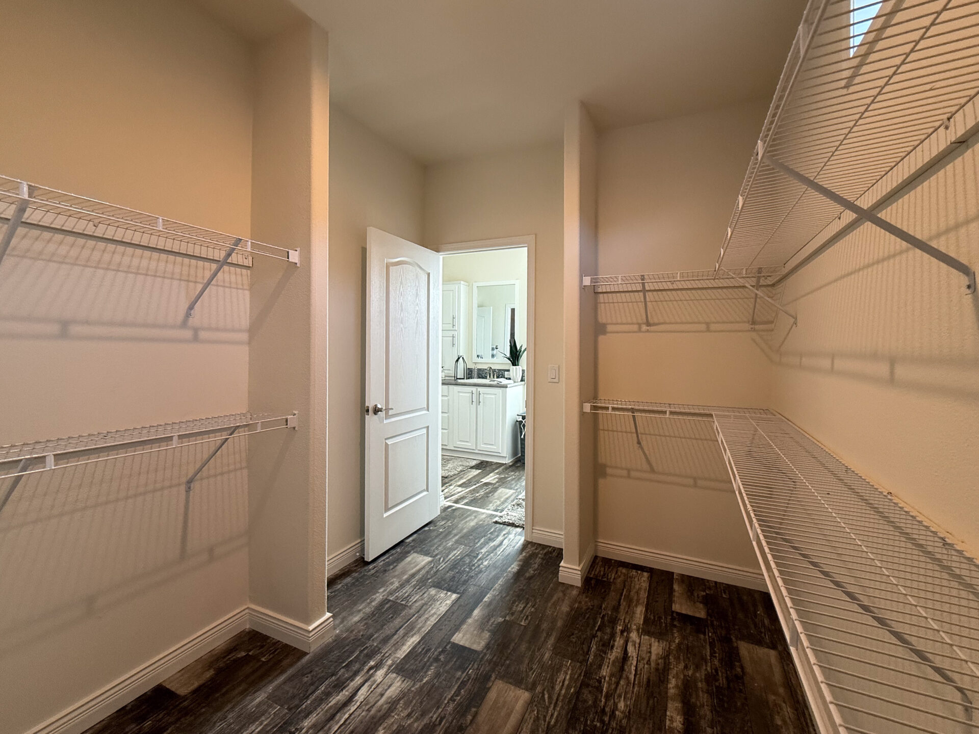 Empty walk-in closet with wire shelving on beige walls, leading to an open white door showing a bright kitchen with wooden floors in the background.