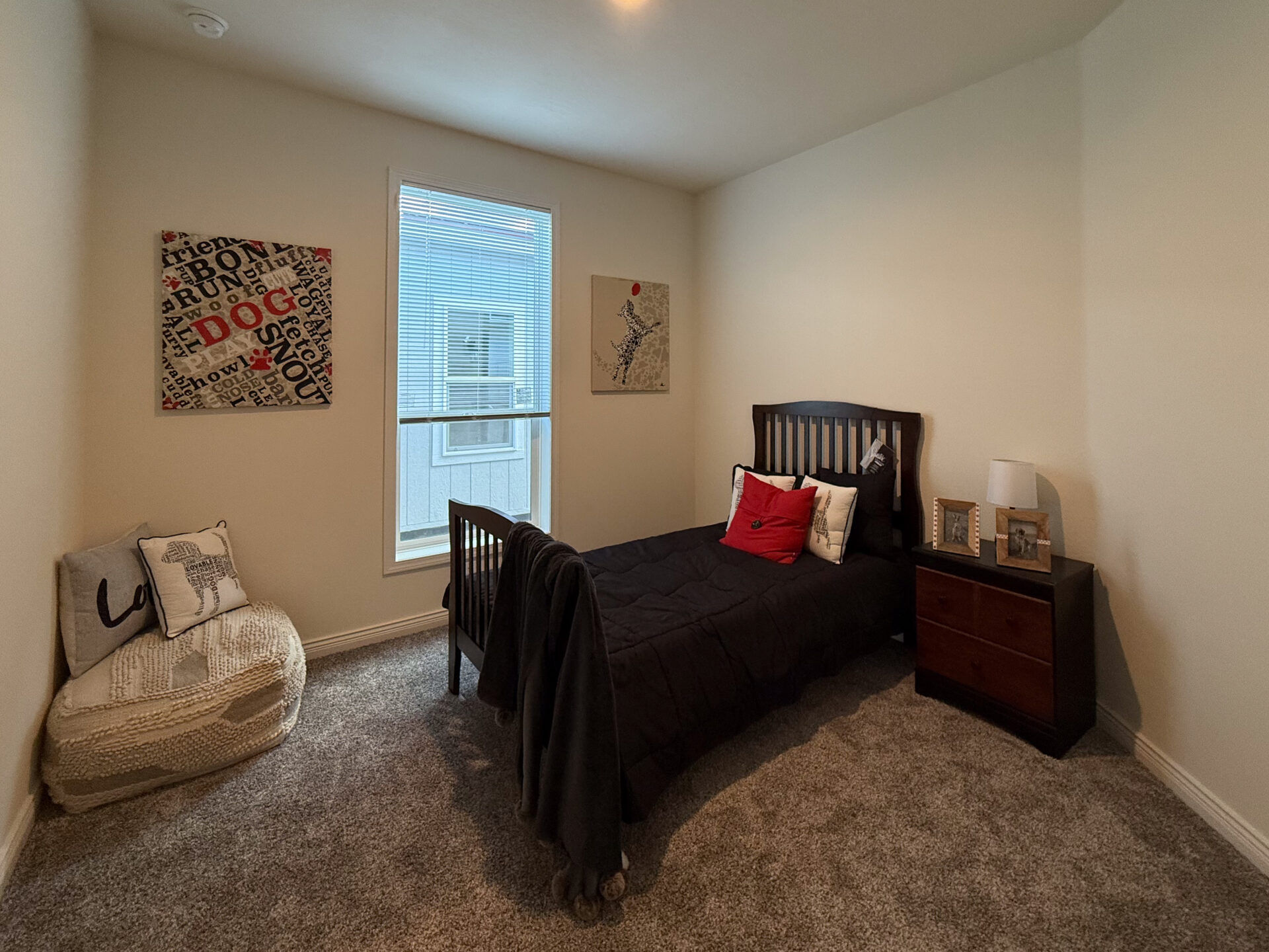 A cozy bedroom with a twin bed covered in a black comforter and red accent pillow. A nightstand with a lamp and framed photos sits beside it. Two modern artworks hang on cream walls. A window with blinds lets in natural light, and a round chair with decorative pillows is in the corner. The room feels warm and inviting.