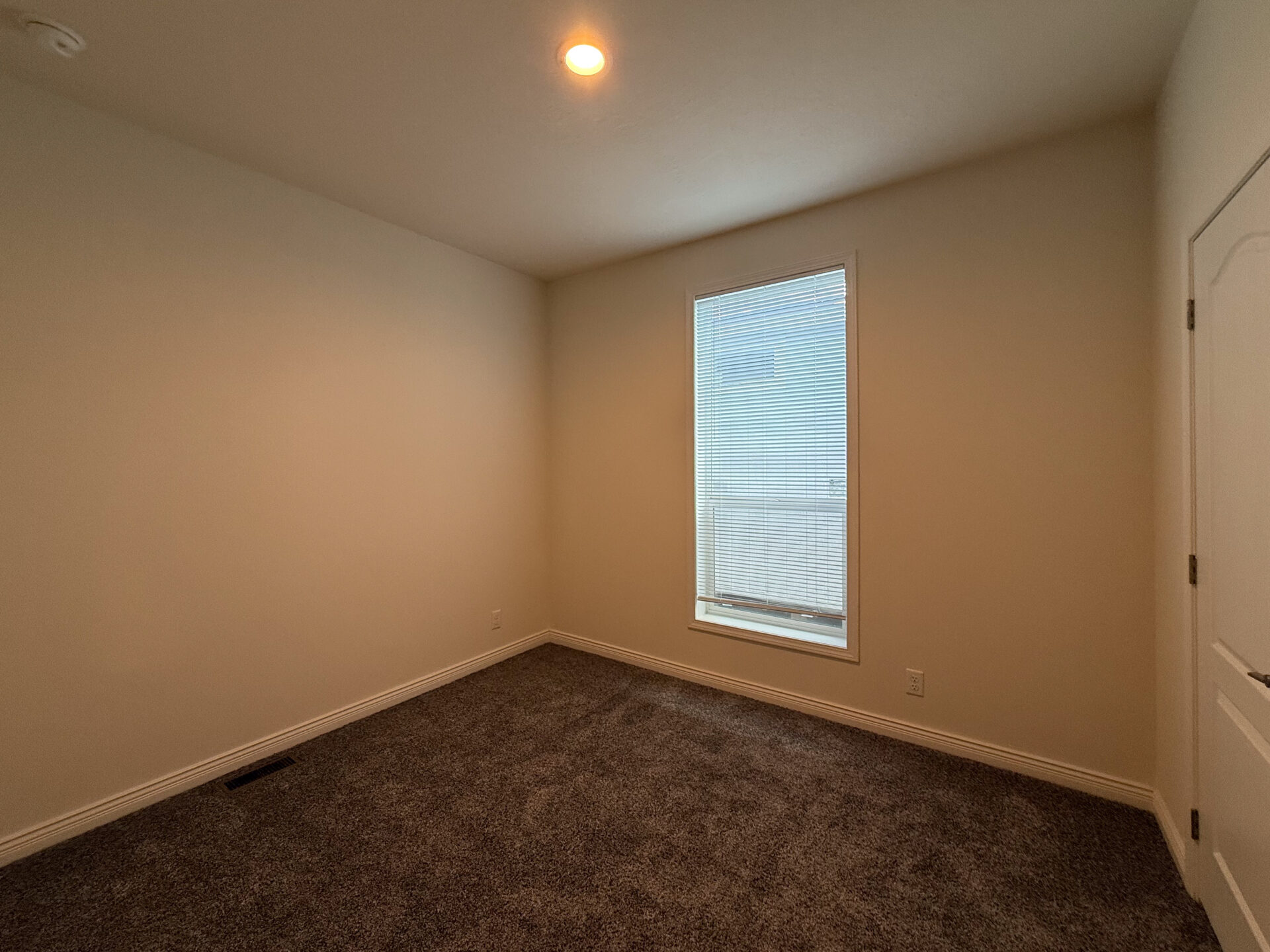 Empty room with beige walls and a single narrow window with closed blinds letting in soft light. Dark carpet flooring and a white door on the right.