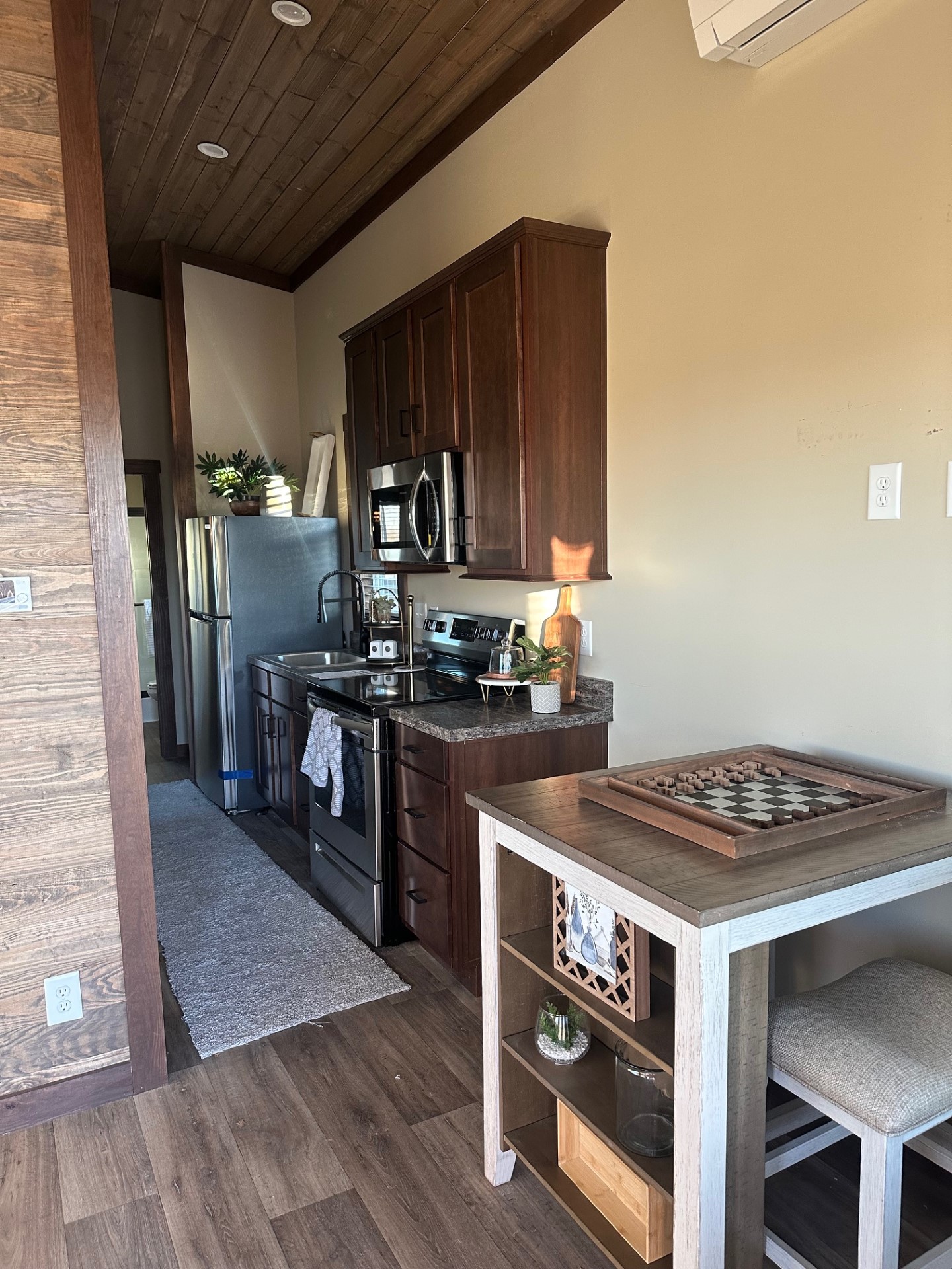 A cozy kitchen with dark wood cabinets and stainless steel appliances. A small table with a checkerboard game and stools adds a welcoming touch.