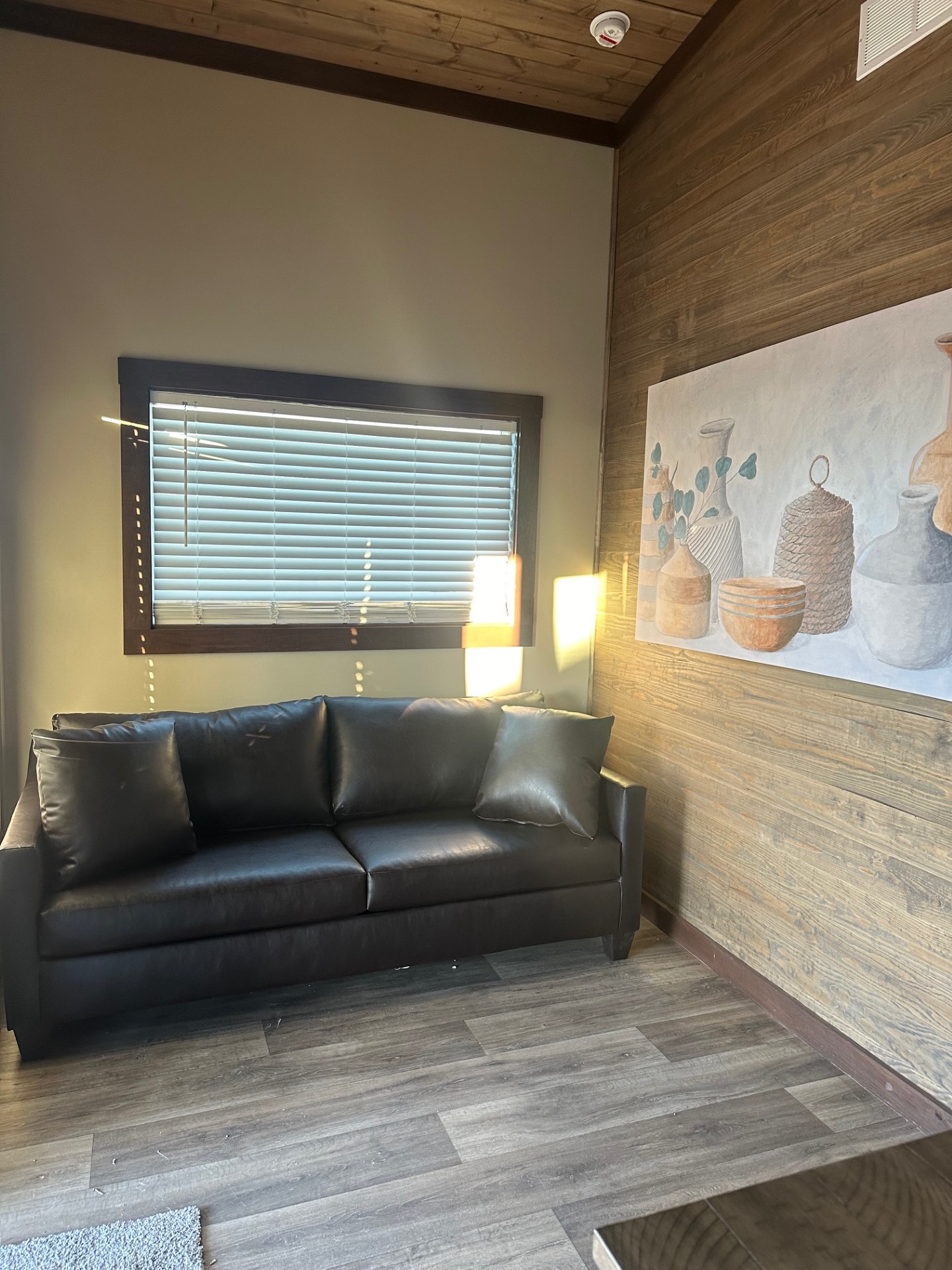 A cozy living room with a black leather sofa, two cushions, wood panel walls, a painting of vases, and a sunny window with blinds, creating a warm atmosphere.