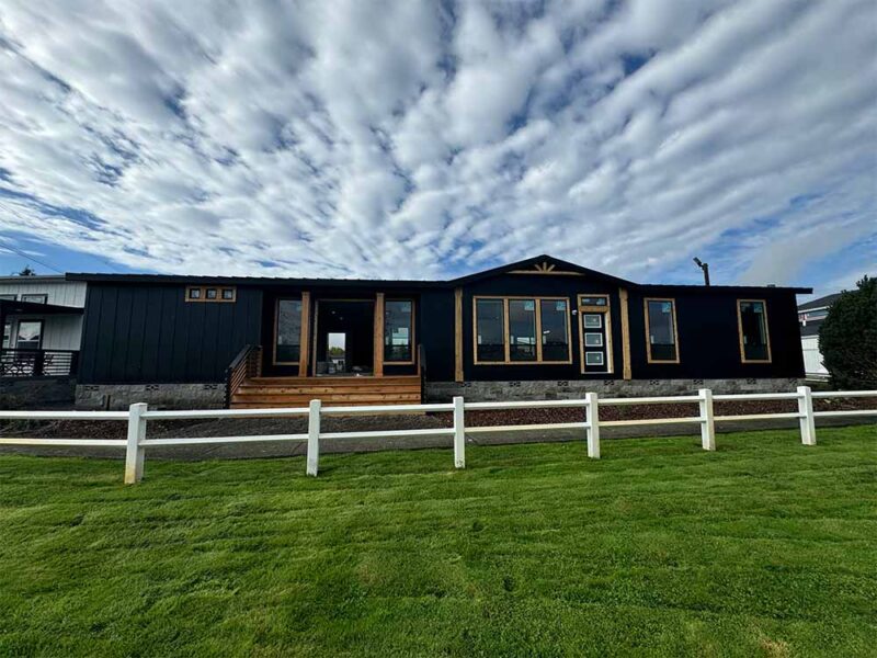 A modern black house with large windows, wooden accents, and a small porch sits behind a white fence on a grassy lawn under a cloudy sky.
