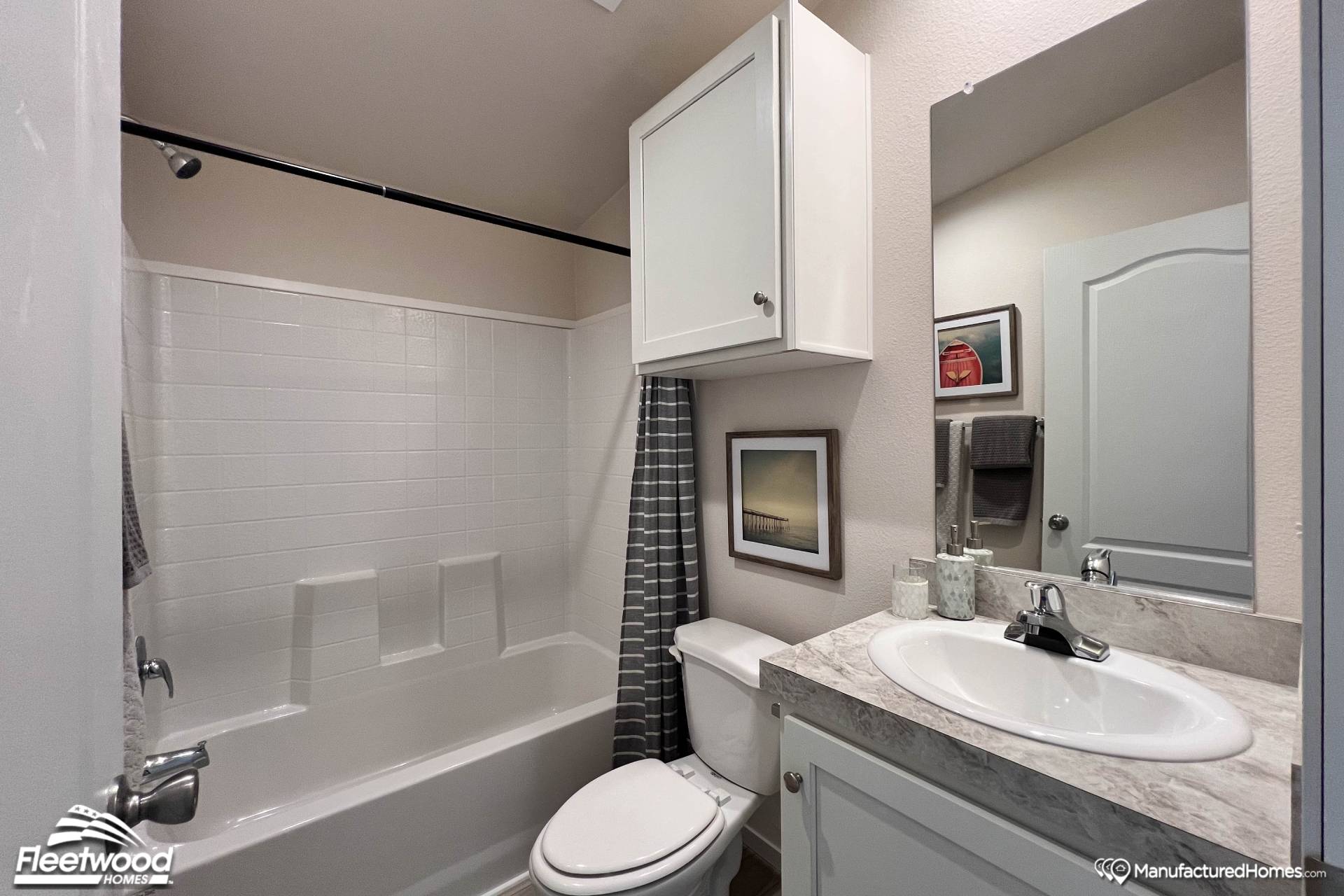 Compact bathroom with white fixtures. Features a bathtub with a gray shower curtain, sink with marble countertop, wall cabinet, and framed art. Modern and tidy.