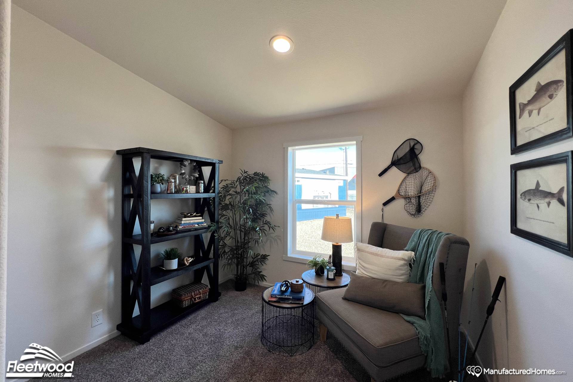 Cozy room with a plush armchair, a small round table, and a tall black bookshelf. Fishing decor hangs on white walls. Soft natural light from a window.