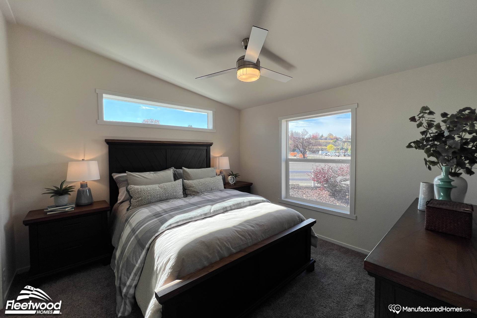 A cozy bedroom featuring a neatly made bed with gray and white linens, flanked by two lamps on wooden nightstands. A ceiling fan and large window let in natural light.