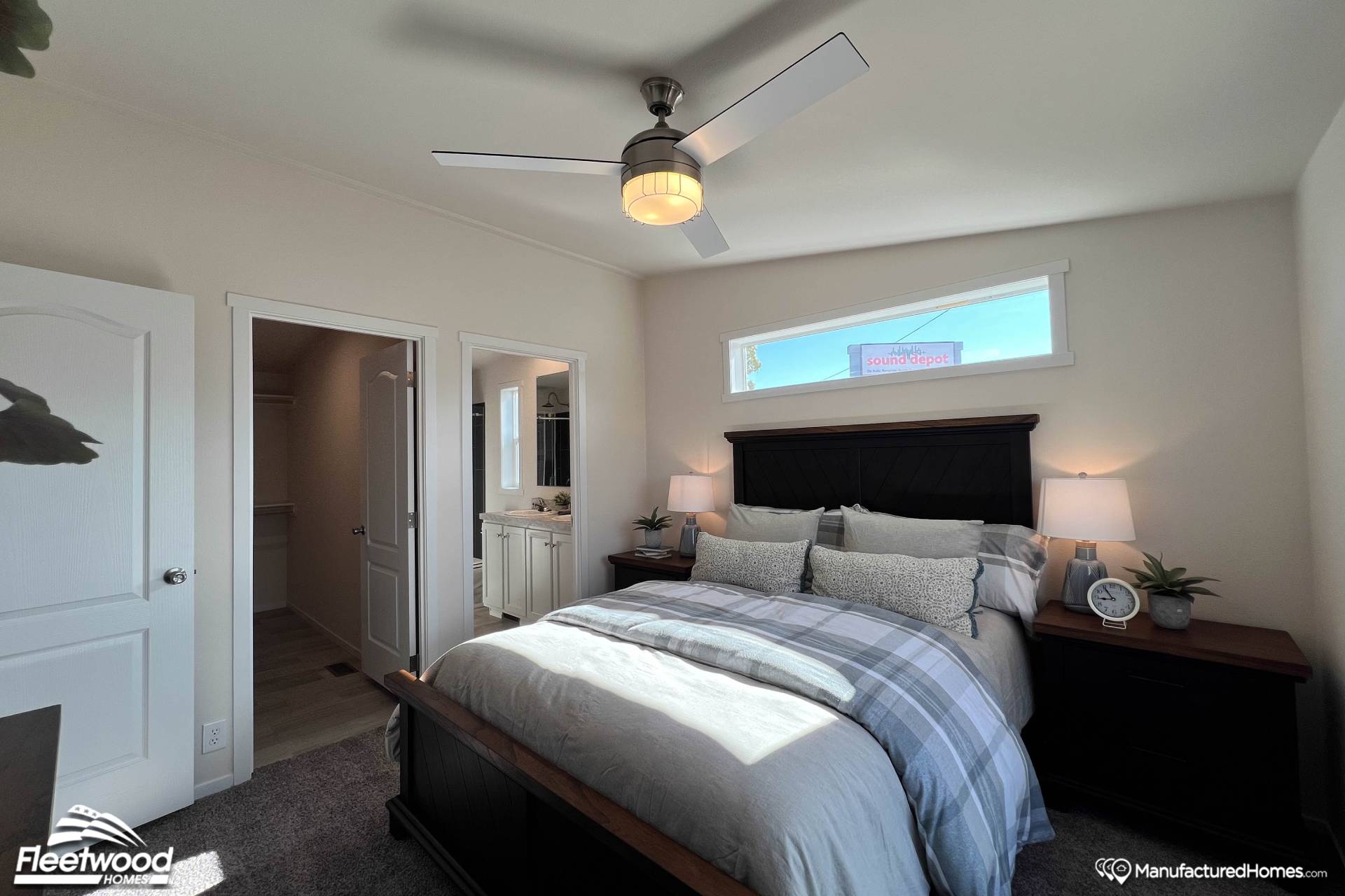 A cozy bedroom features a neatly made bed with plaid bedding, flanked by wooden nightstands. A modern ceiling fan hangs overhead, and sunlight streams through a small window, creating a peaceful ambiance. A door leads to an adjoining bathroom.