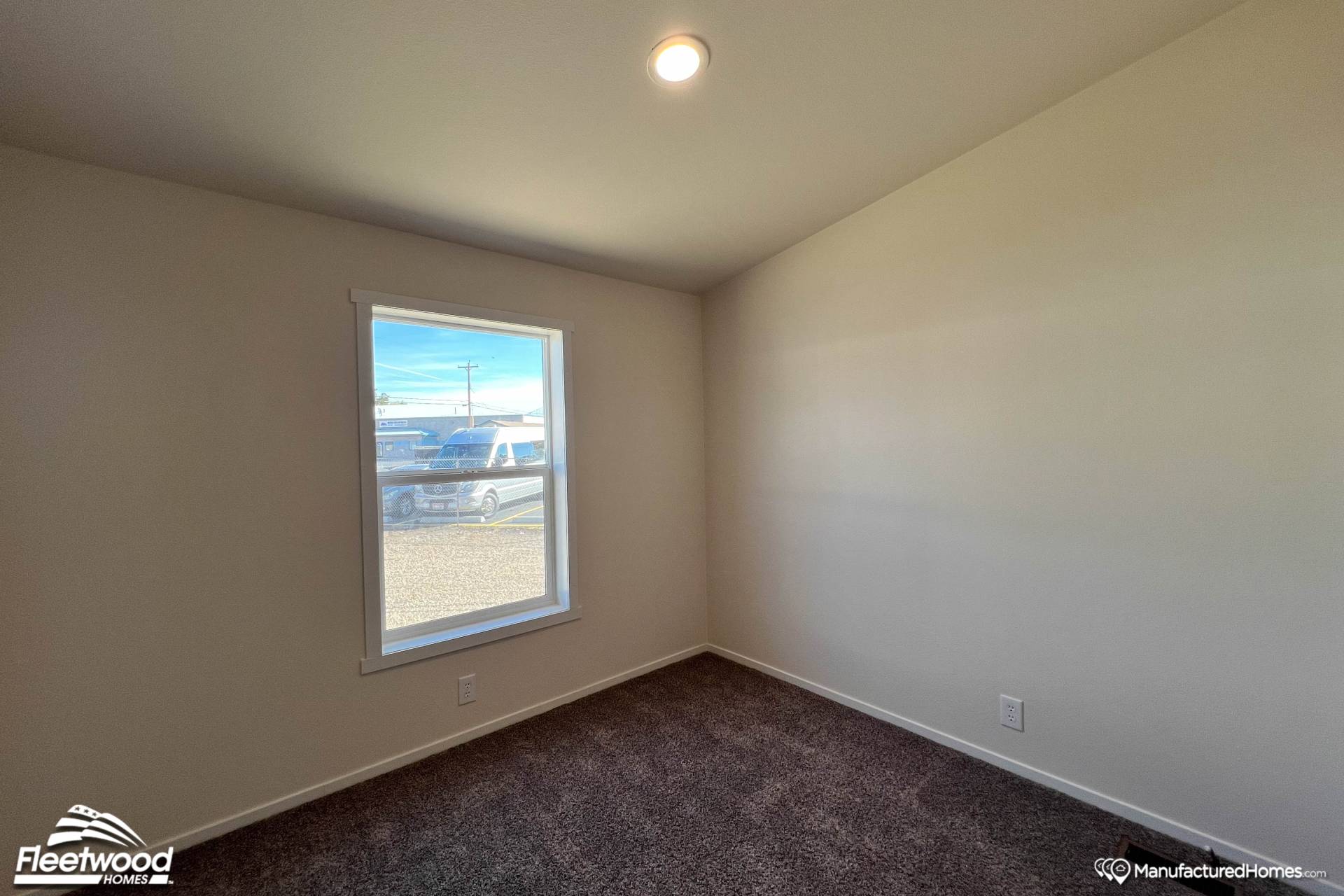 Small, empty room with beige walls and a dark carpeted floor. A single window reveals parked cars and a clear blue sky outside. Soft overhead lighting.