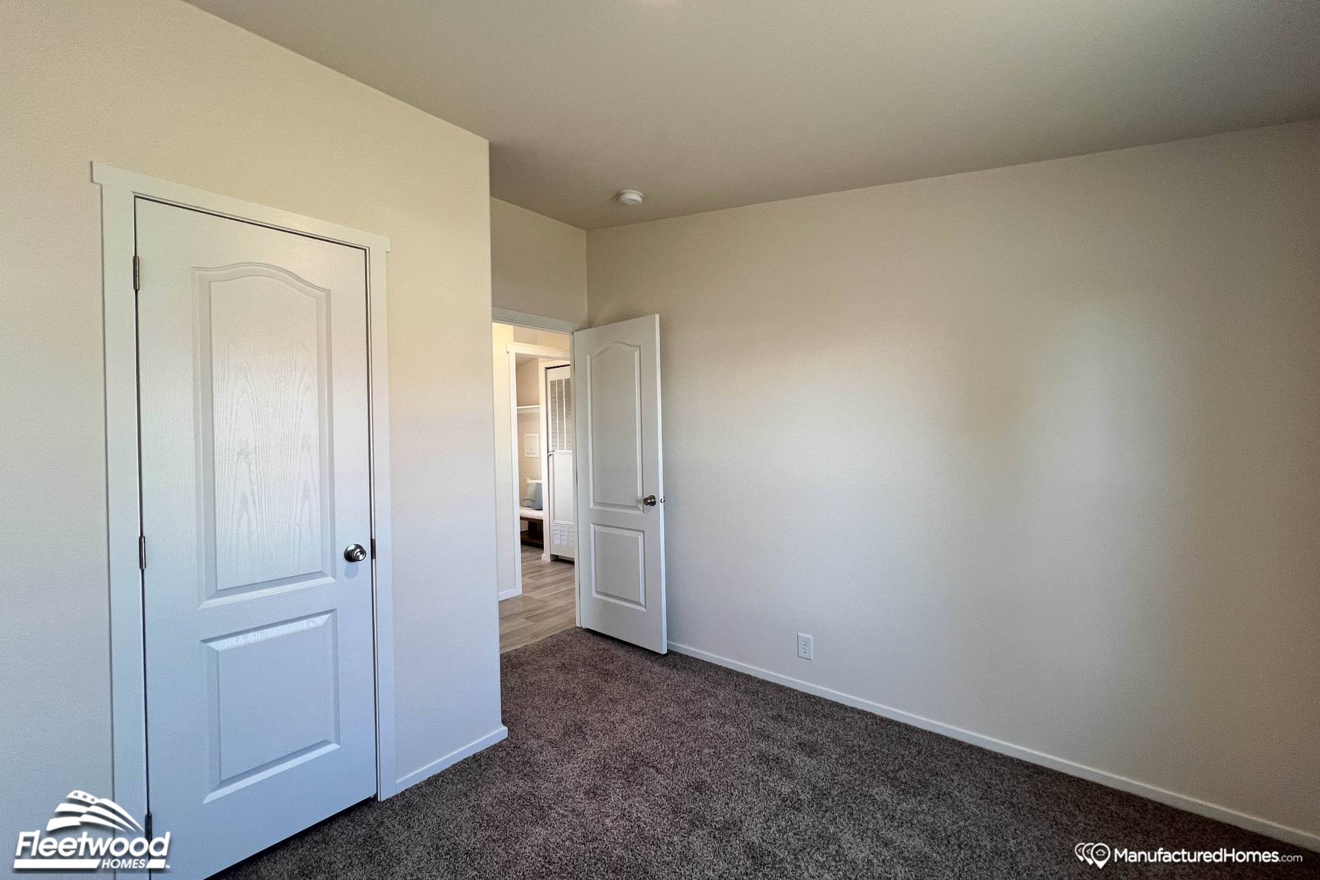 Empty, carpeted room with beige walls, two white doors, and soft natural light. The space feels clean, simple, and open, suggesting a new living area.
