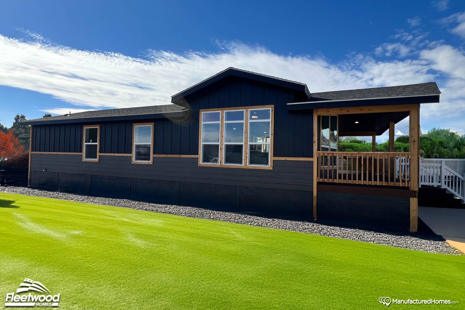 A modern black manufactured home with large windows and a front porch sits under a clear blue sky. It is surrounded by a neatly maintained green lawn.