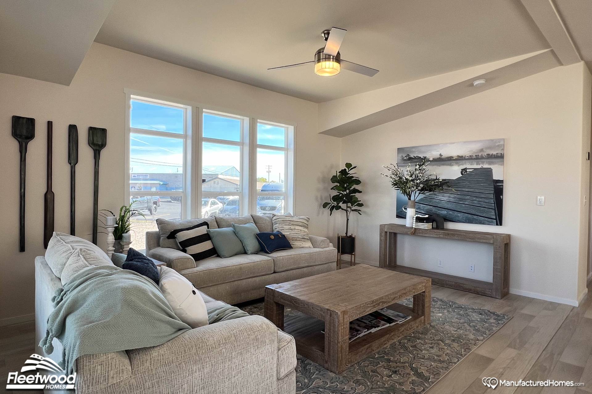 A cozy living room with neutral tones features two beige sofas adorned with pillows and blankets, a wooden coffee table, and decorative oars on the wall. Large windows brighten the space, revealing a serene outdoor view. A ceiling fan hangs above, and a photograph of a dock complements the room's modern decor.
