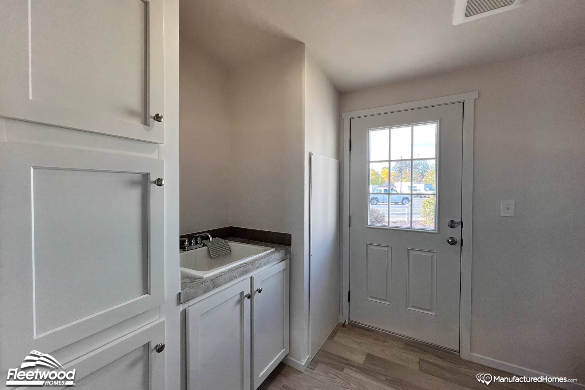 A bright, compact laundry room with white cabinets, a sink, and a door with glass panels showing a sunny outdoor scene. Warm and functional space.