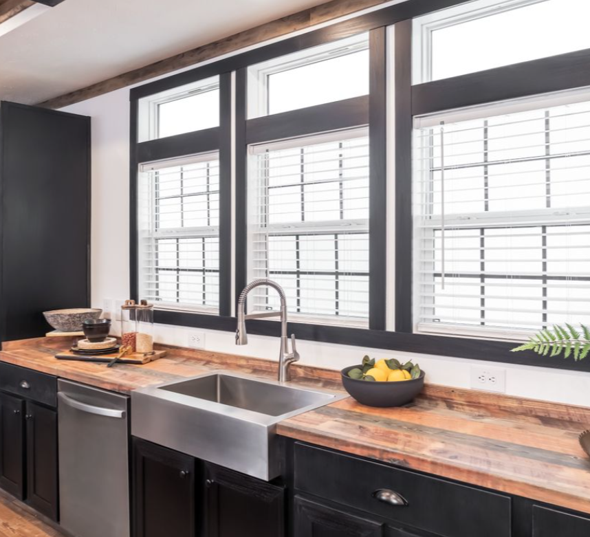 A modern kitchen with a large farmhouse sink, wood countertops, and black cabinets. Three large windows let in light, creating an inviting atmosphere.