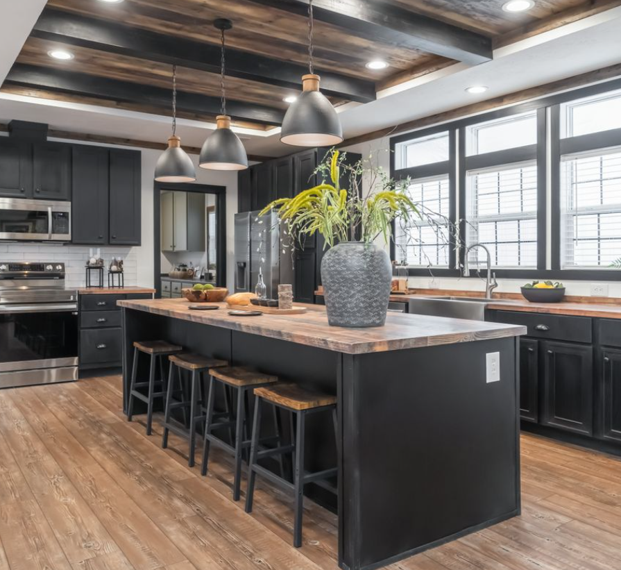 Chic kitchen with dark cabinets, wood accents, and large island featuring bar stools. Pendant lights hang above, and sunlight streams through windows.
