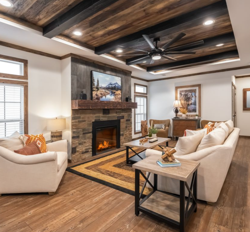 Cozy living room with rustic decor, featuring a stone fireplace, white sofas with orange cushions, and a wooden coffee table on a patterned rug. Wood beam ceiling adds warmth.