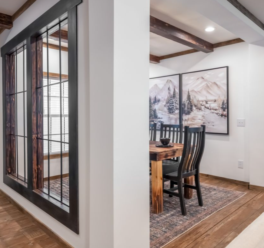 A cozy dining area with a wooden table and chairs, mountain landscape paintings, and rustic wood beams. A large window with dark frames adds warmth.