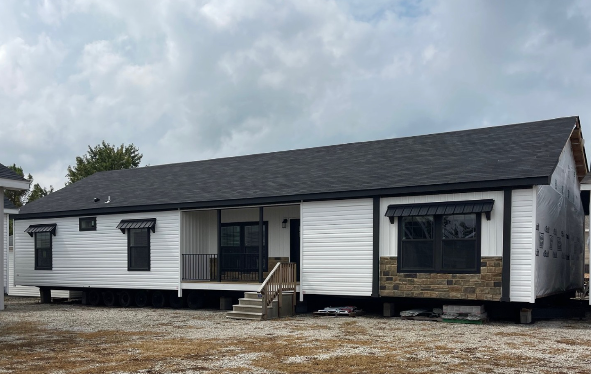 A long, modern single-story house with white siding and a dark roof. Stone accents adorn the exterior. The sky is overcast, suggesting a calm, subdued atmosphere.
