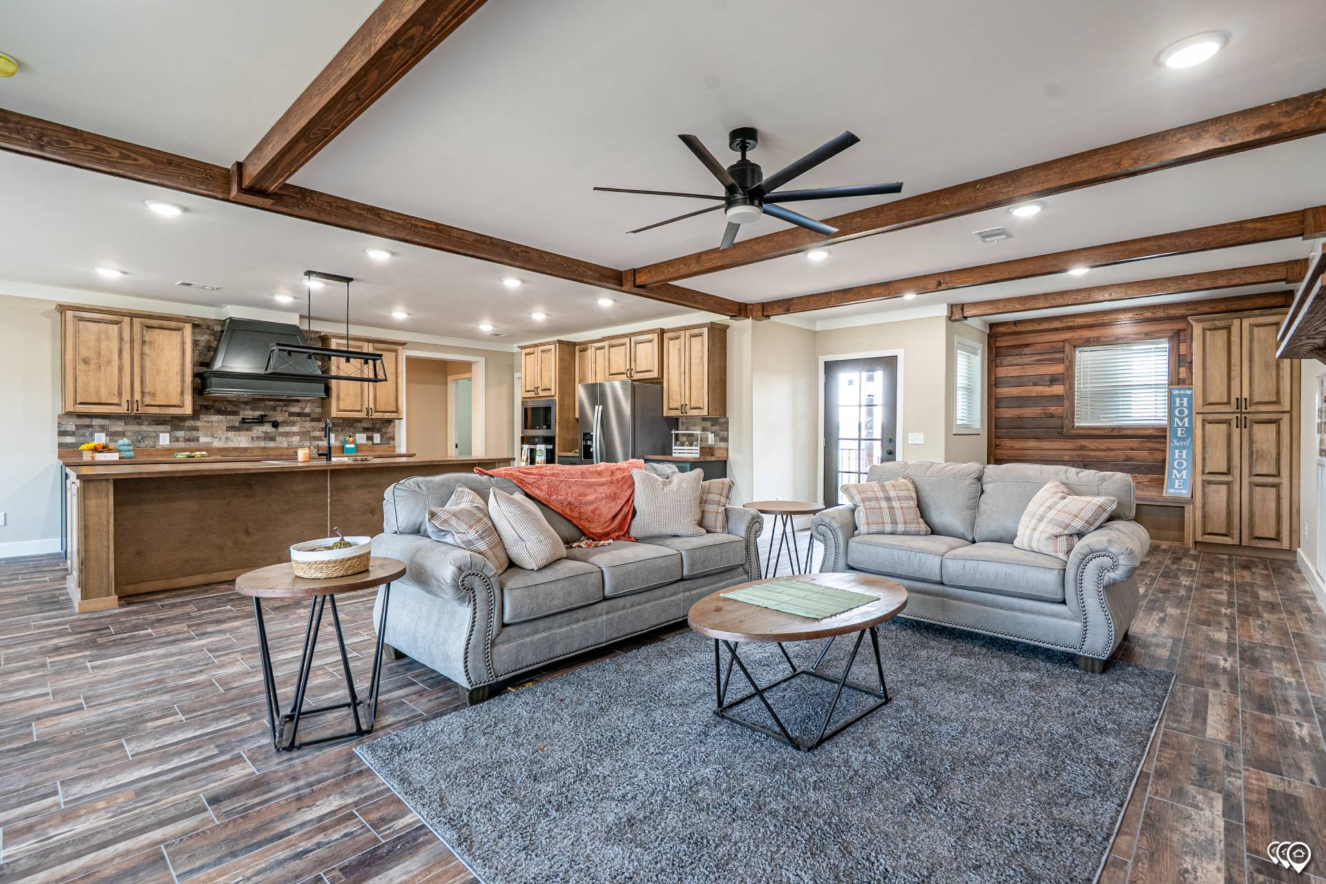 Spacious living room with gray sofas, wooden beams, and a ceiling fan. Cozy ambiance with an open kitchen featuring wooden cabinets. Warm tones.