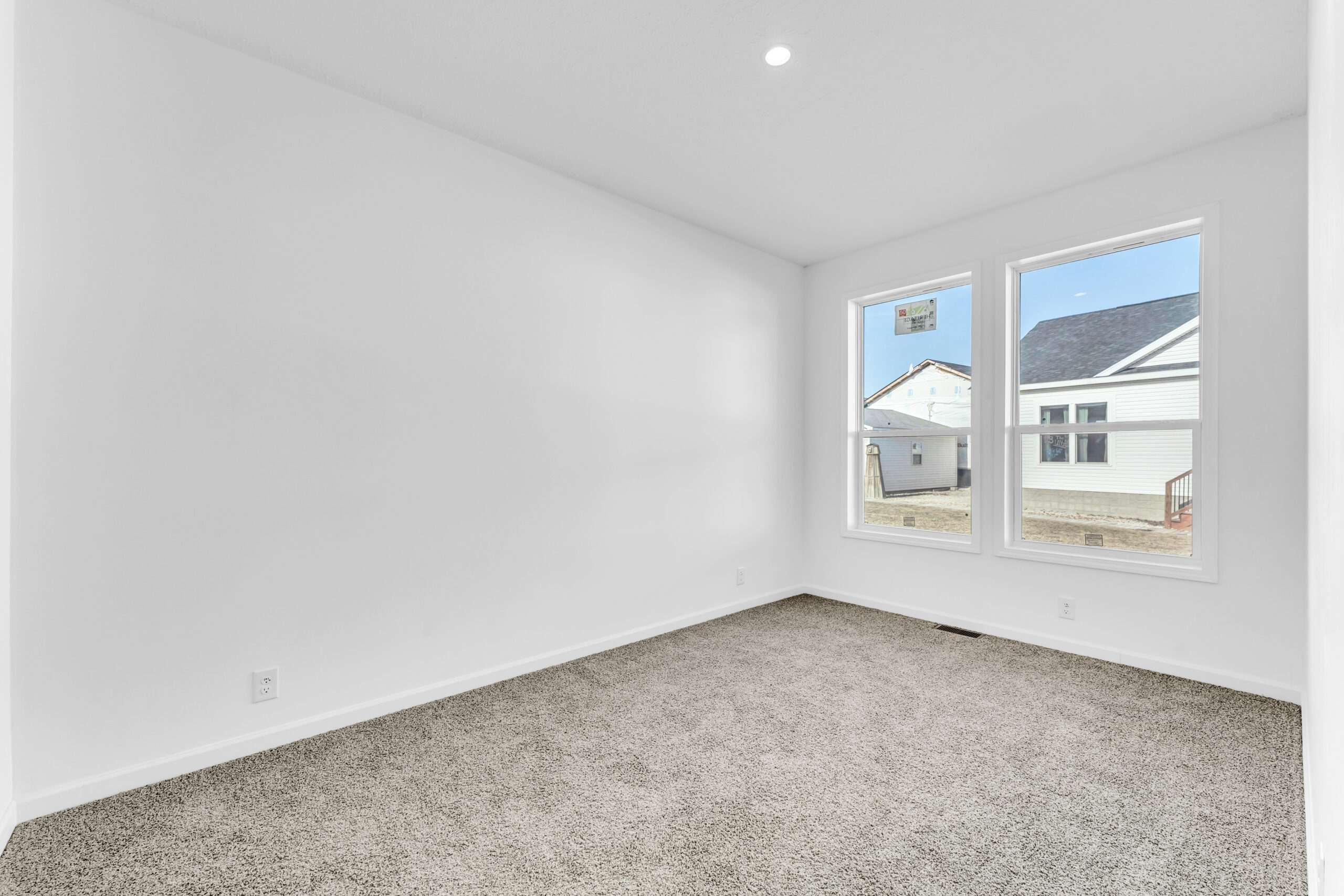 Empty room with white walls, a beige carpet, and two large windows showing an outdoor view of neighboring houses under a clear blue sky. Bright and minimalistic.