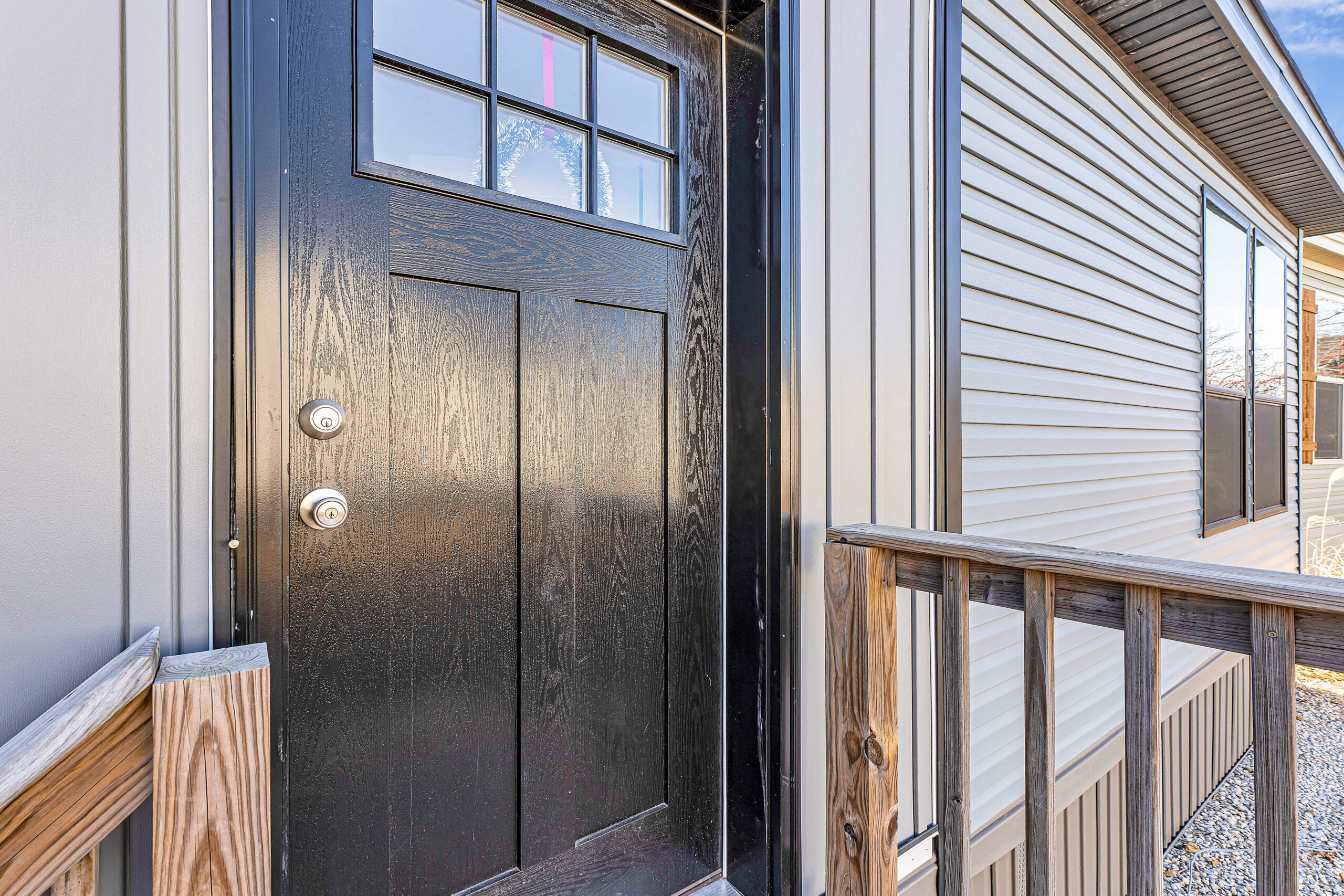A modern black front door with small windows, flanked by beige vinyl siding. It has a wooden porch railing, conveying a welcoming, contemporary feel.