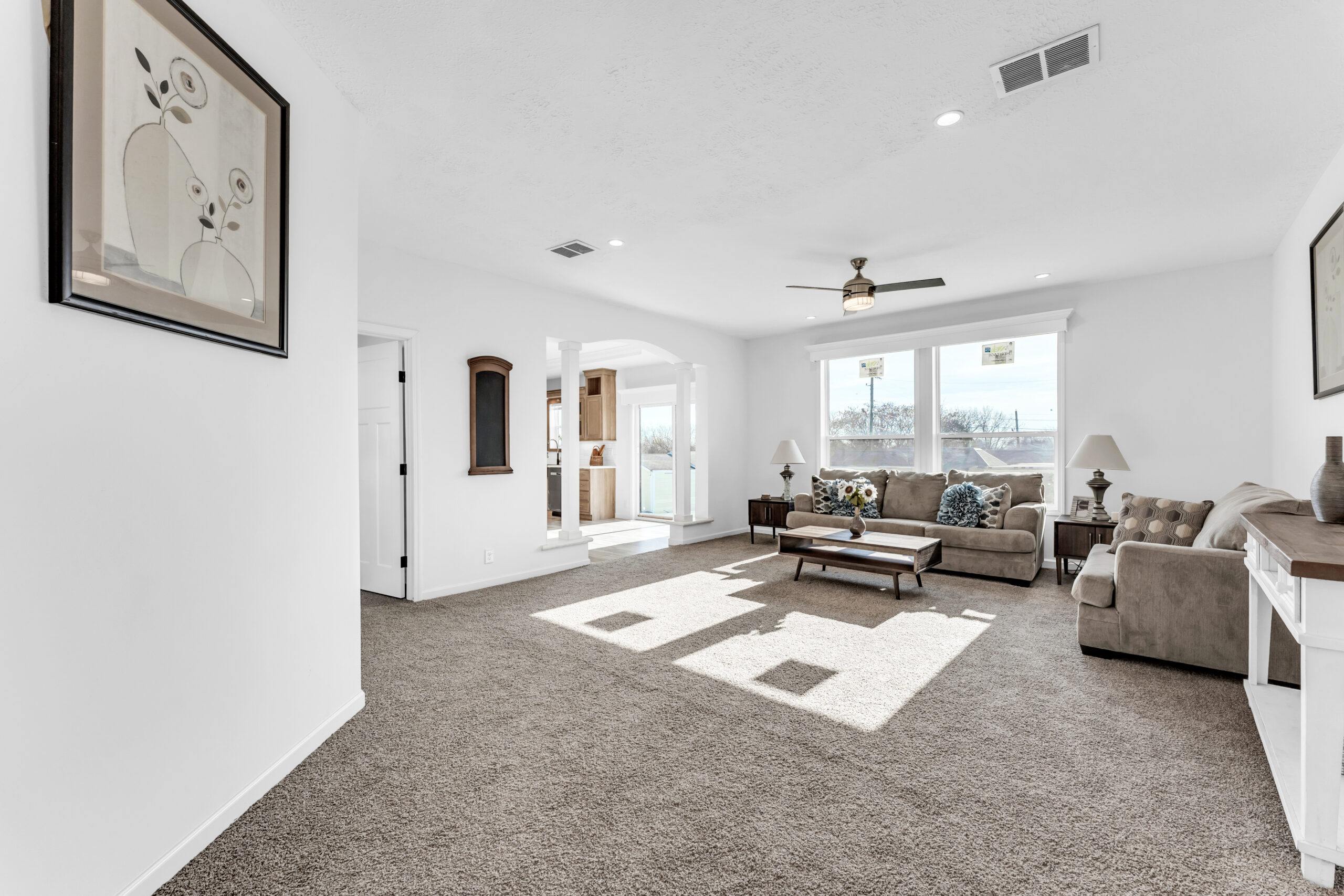 Bright living room with plush gray carpet, beige sofas, and colorful cushions. Large windows bring in natural light. Minimalist decor with framed art.