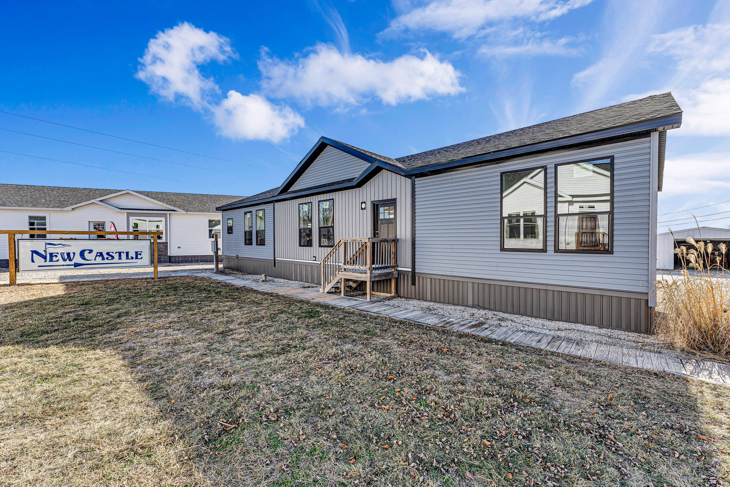 Single-story modular home with gray siding and black roof. A sign reads "New Castle" on the lawn. The sky is bright blue with clouds, creating a welcoming atmosphere.