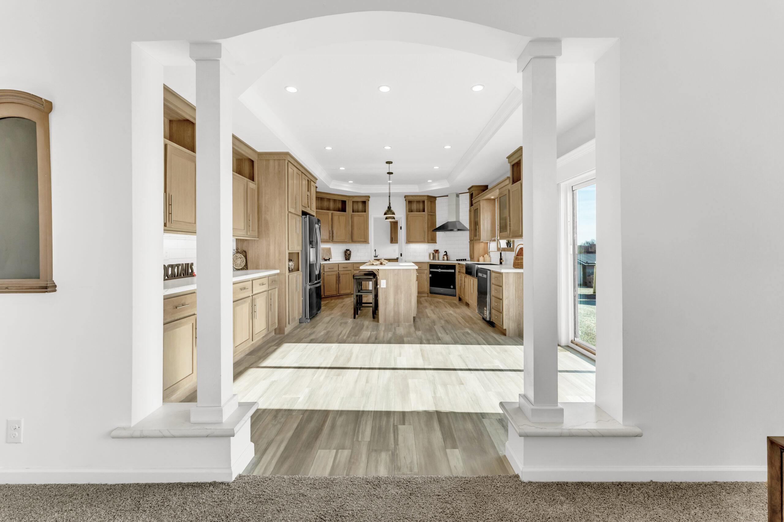 Spacious kitchen view through an arched doorway features light wood cabinets, a central island with bar stools, and stainless steel appliances.